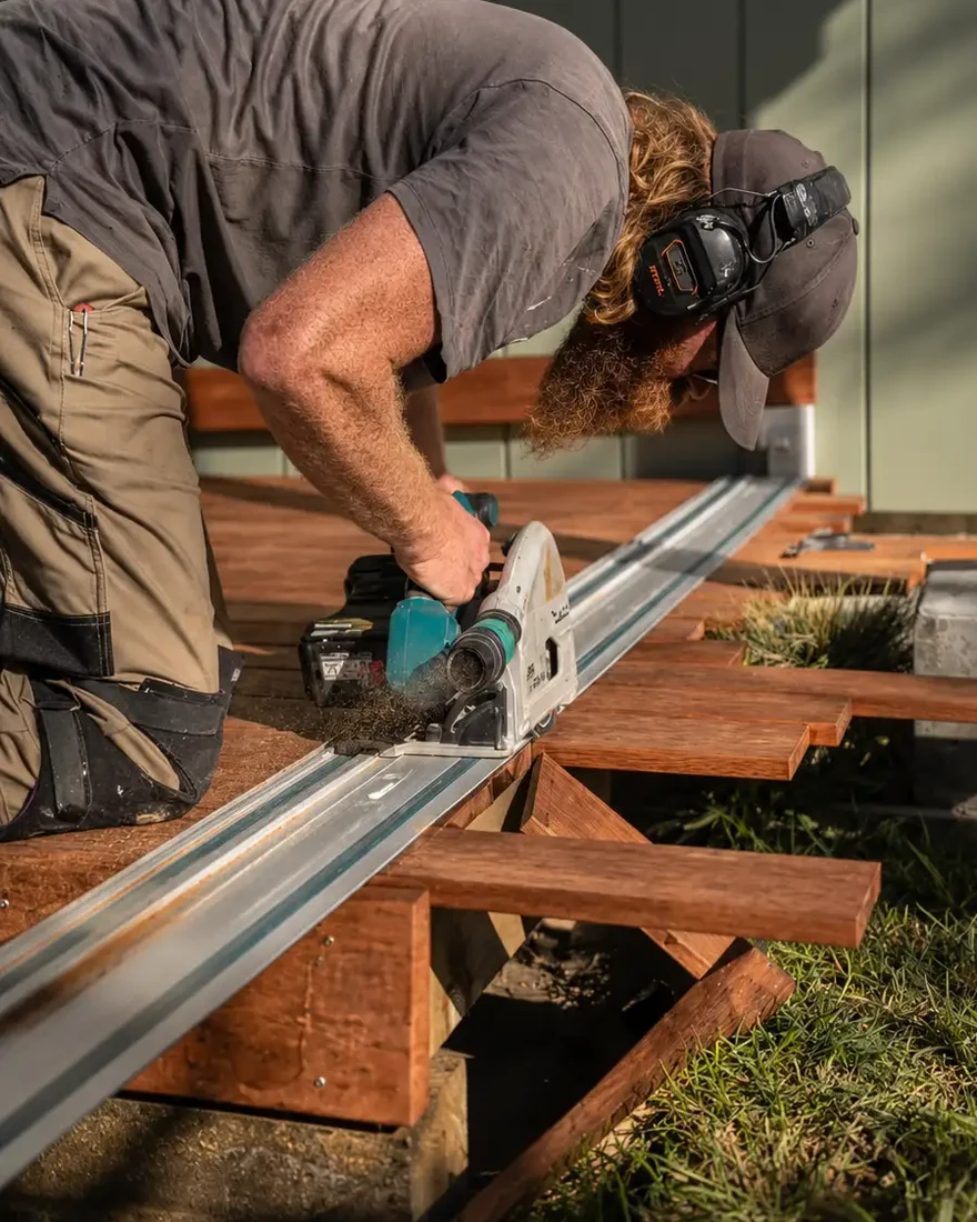 Clinton bent over a track saw, cutting a decking board on site in late afternoon light.
