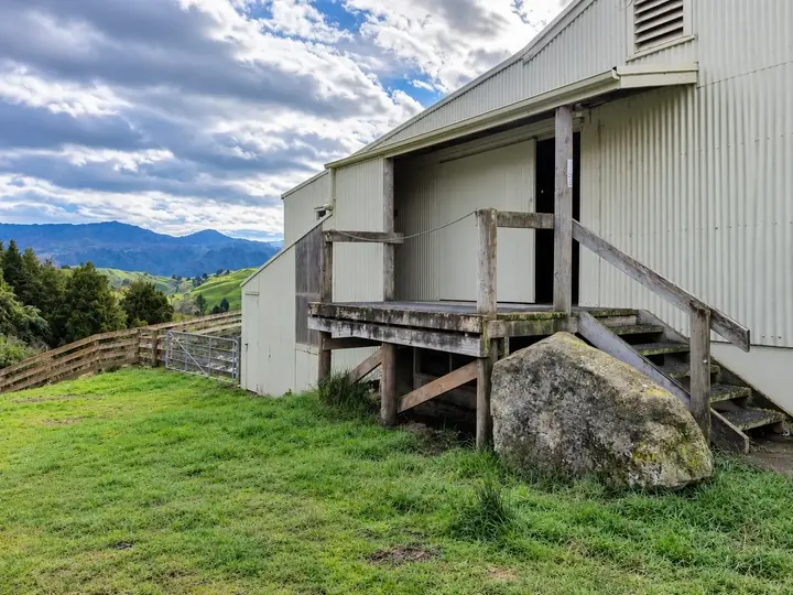 Taumarunui Woolshed — the end of the shed from the paddock, its timber loading platform standing off the corrugated wall with the hill country behind