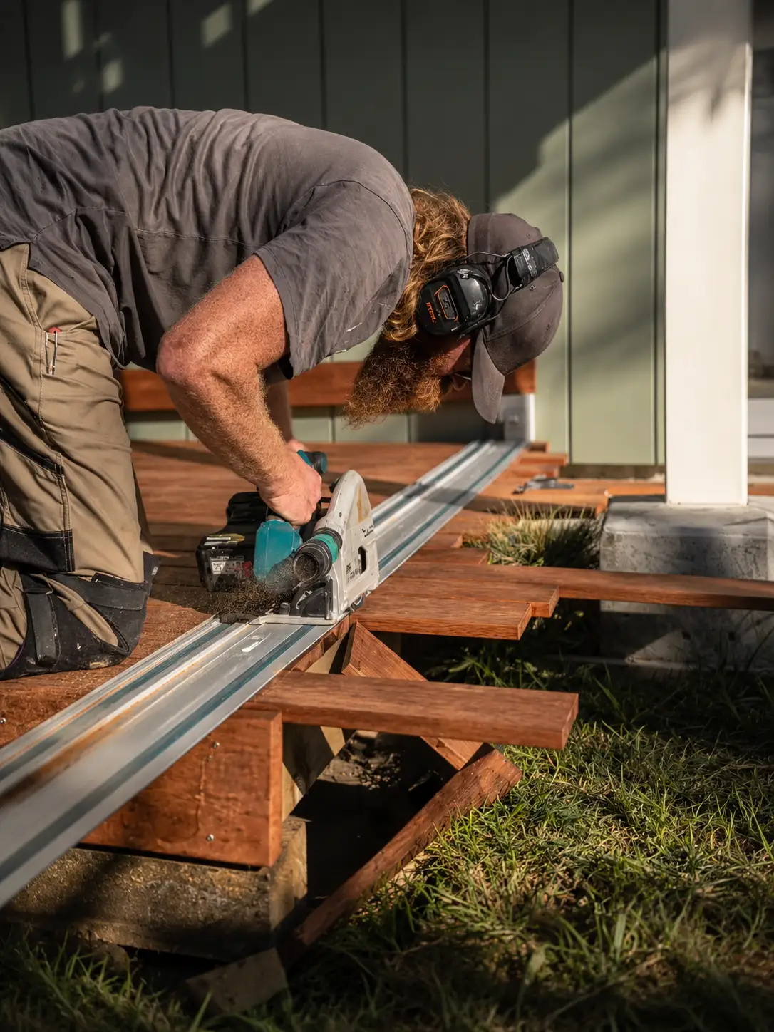 Clinton bent over a track saw, cutting a decking board on site in late afternoon light.