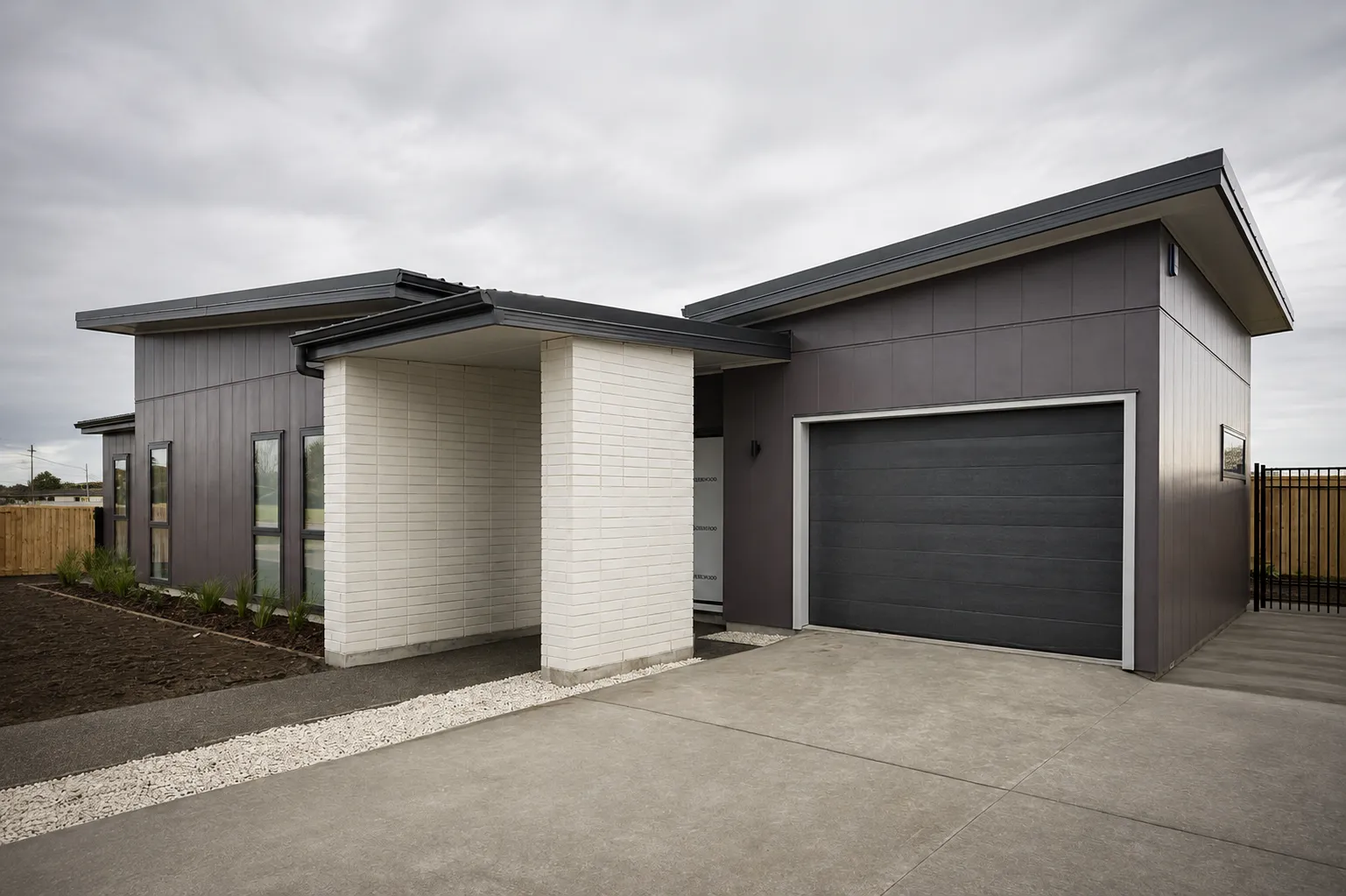 Ōtaki Gardens — the finished showhome from the driveway, dark vertical cladding and a pale brick entry portico under a mono-pitch roof