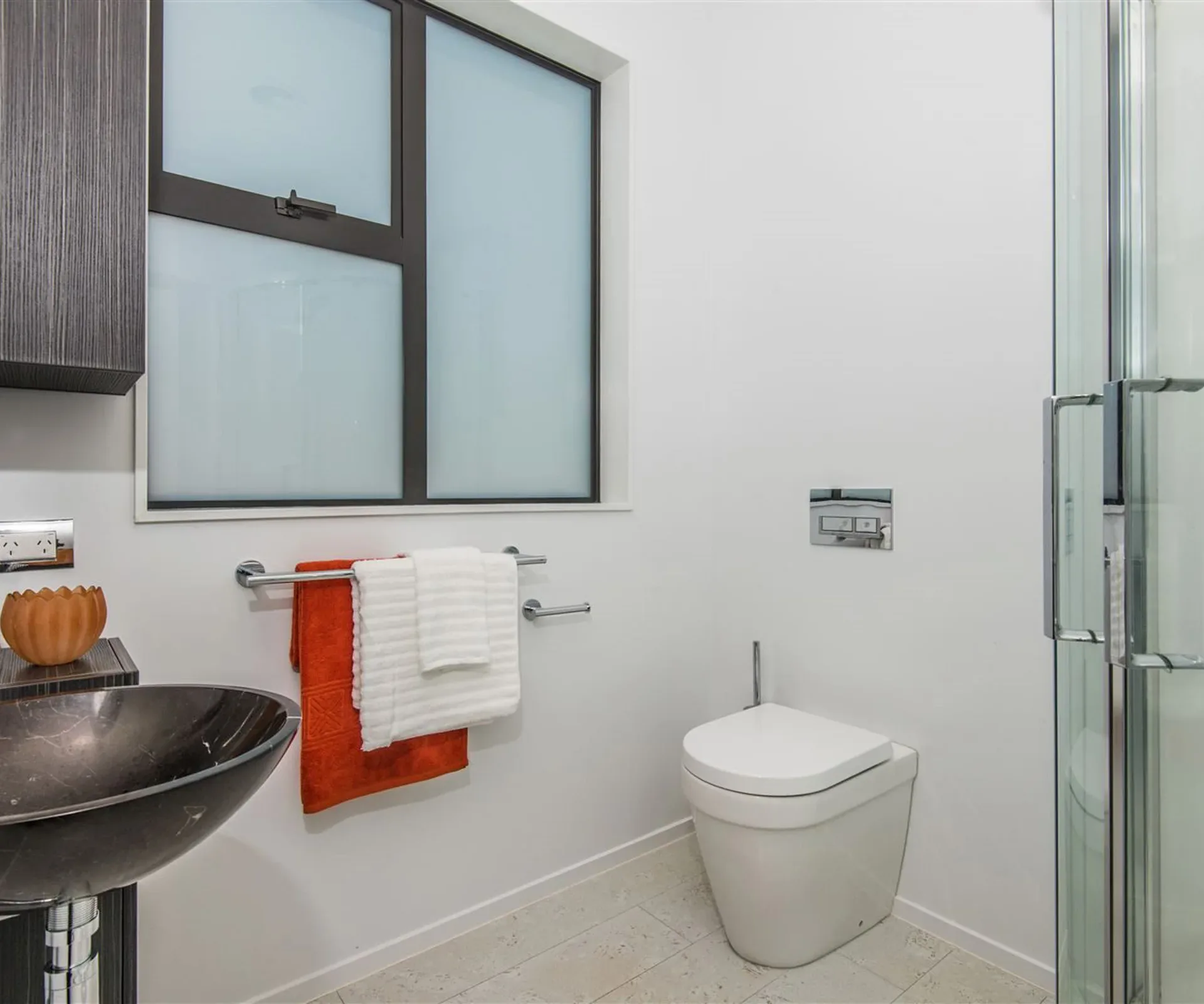 A bathroom — a dark stone vessel basin on a timber vanity, a wall-hung toilet, and the shower behind glass under an angled obscure-glazed window.
