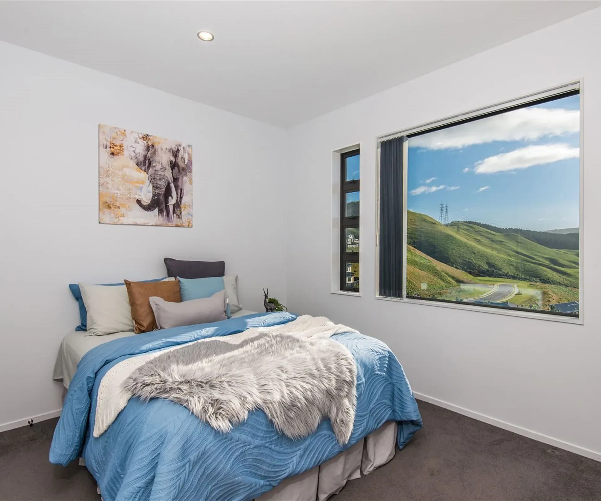 A bedroom in blue — the bed against the wall under a painted elephant canvas, and a wide window onto the green hills and the road running below them.