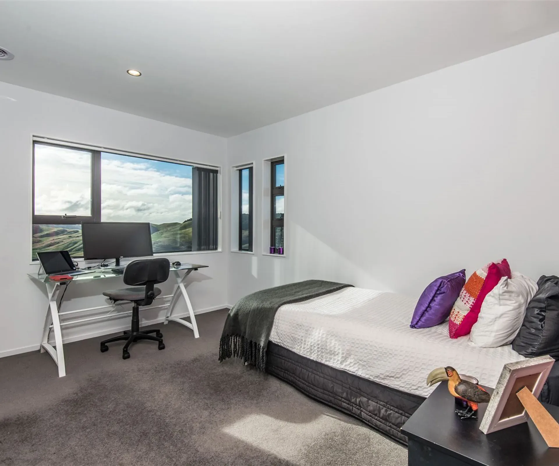 A bedroom set up as a study — a glass desk with a monitor and office chair under the corner windows, the bed alongside it, and the hills filling both runs of glass.