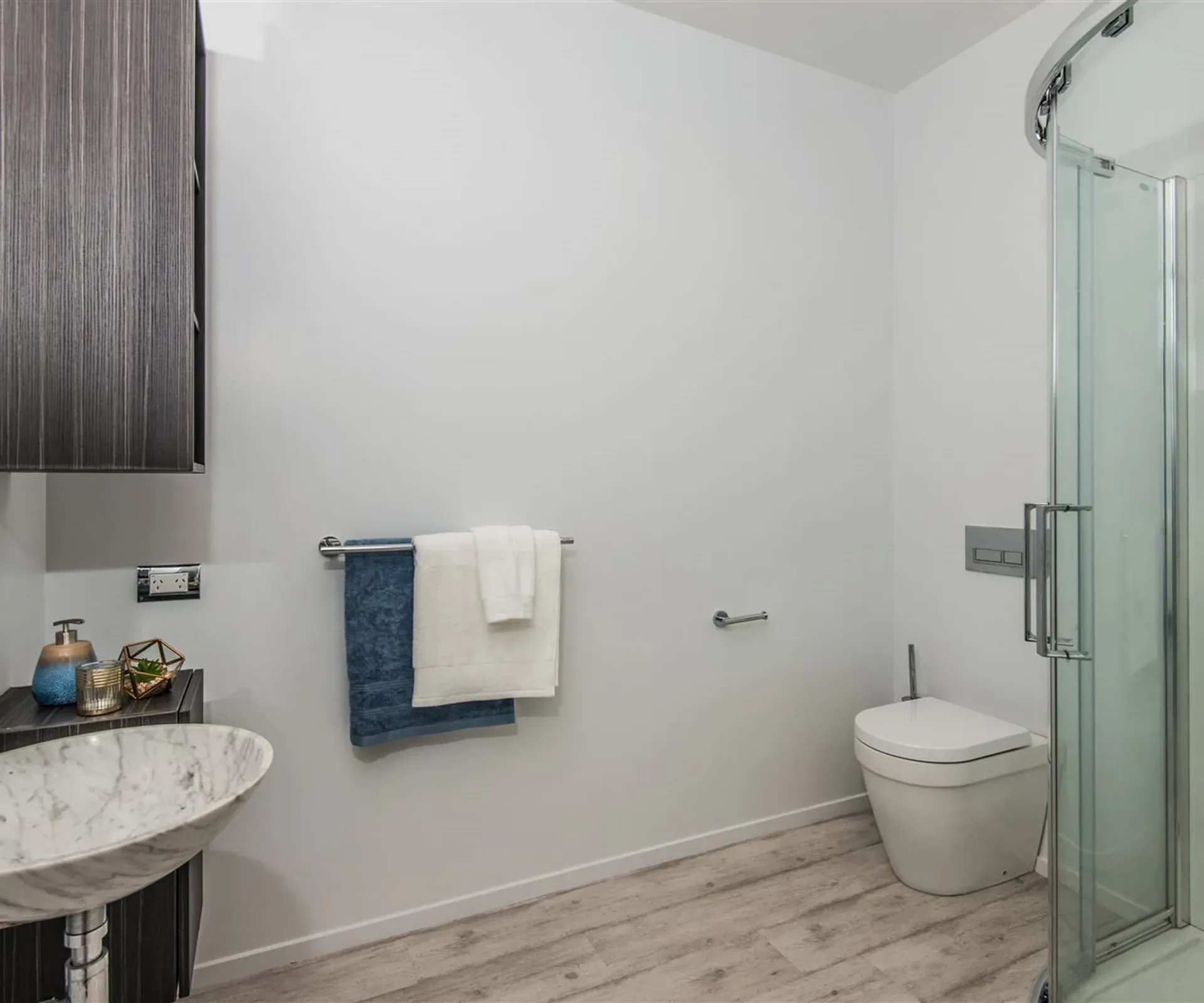 The family bathroom — a marble vessel basin on a dark timber vanity, a wall-hung toilet, and a curved glass shower over timber-look flooring.