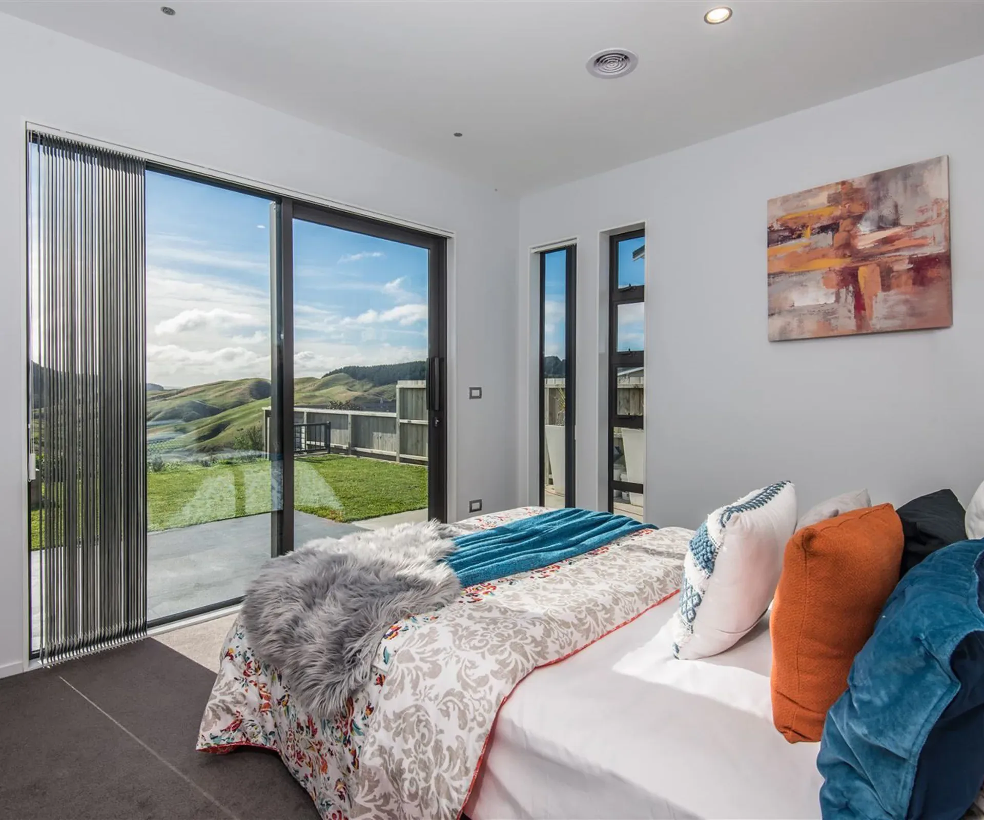 A ground-floor bedroom — the bed beside a full-height slider standing open onto the paving and lawn, with the green hills beyond the fence.