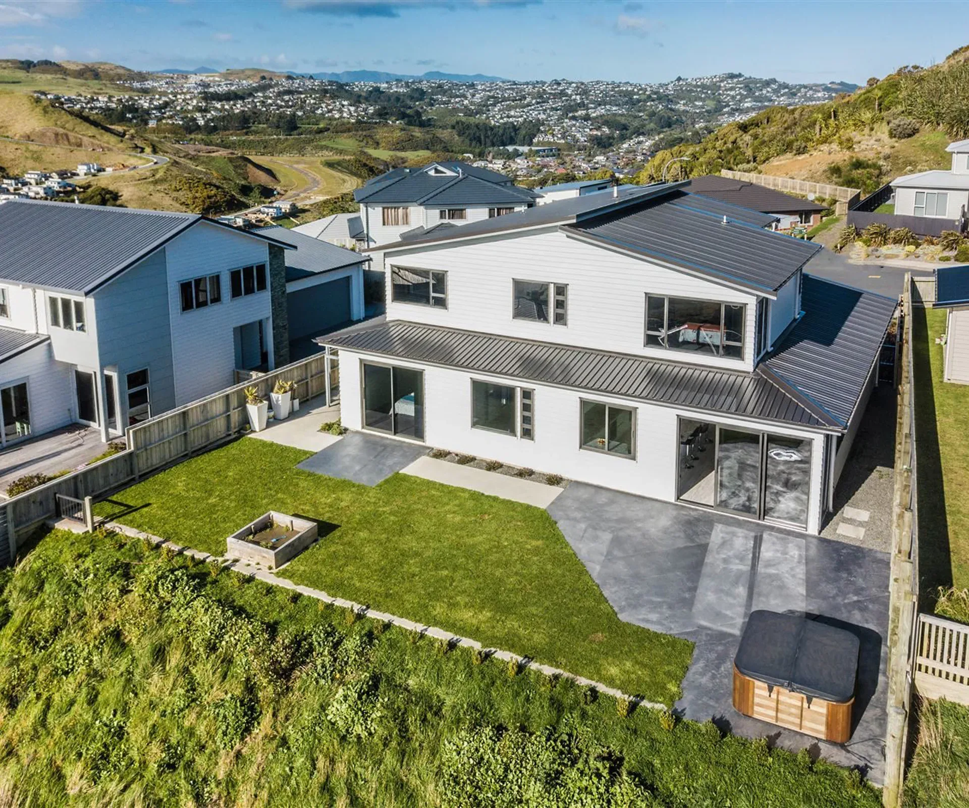 The house from above — the roof form and the rear lawn with its raised garden bed, the neighbouring houses along the street, and the valley and hills beyond.