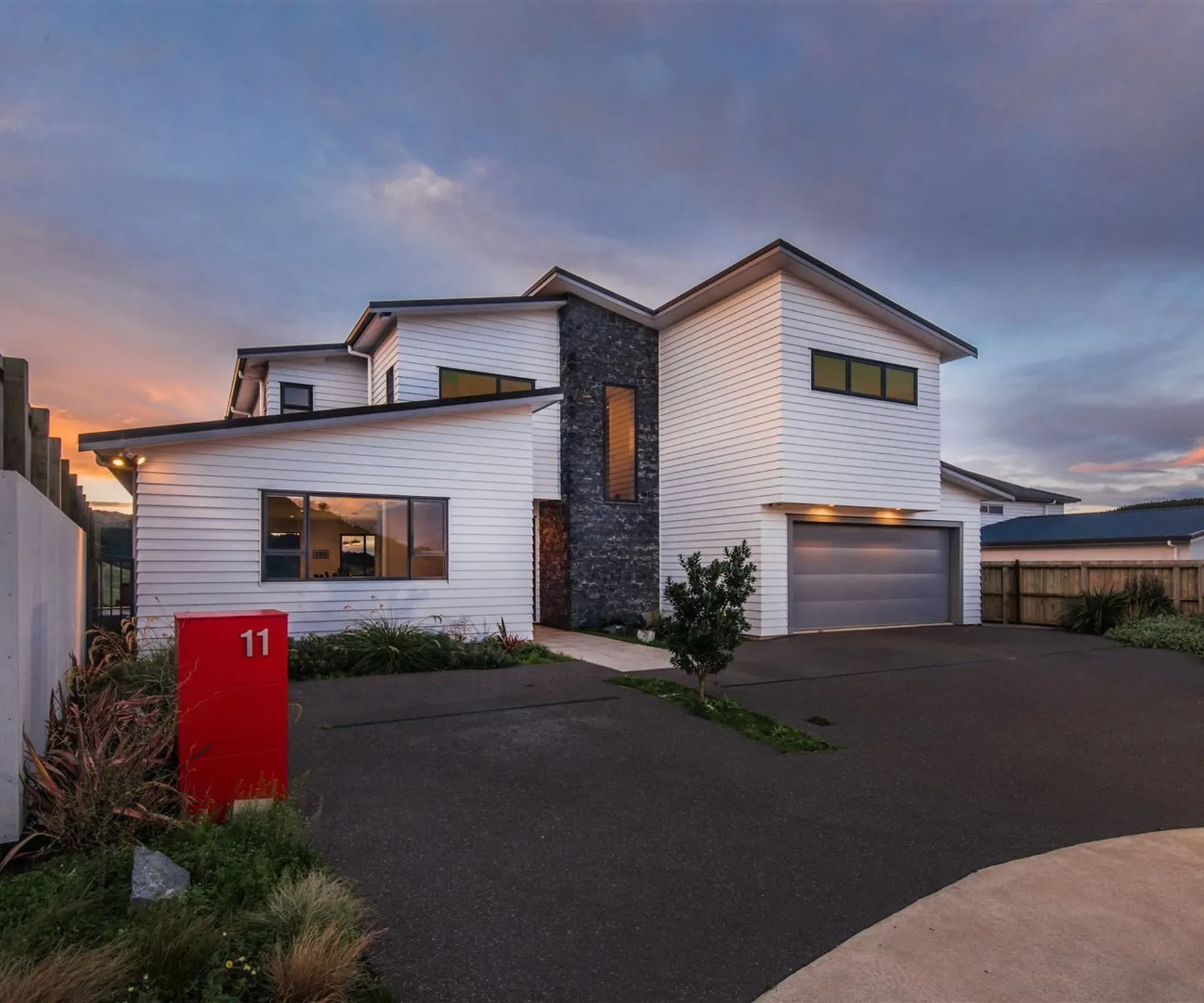 The house from the driveway at dusk — the white weatherboard elevations stepping around the dark schist entry column, the garage lit from above, and the red letterbox at the kerb.