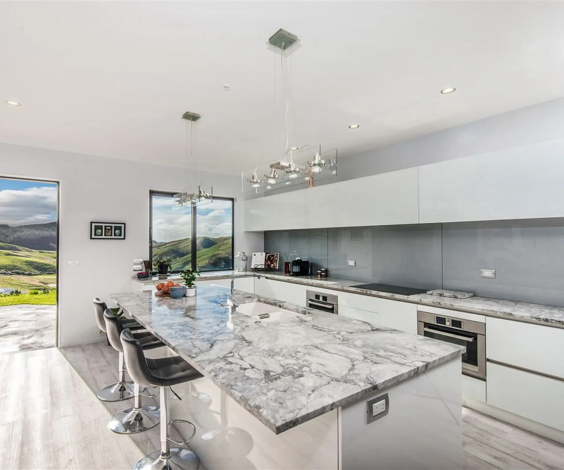The kitchen — a marble-topped island with three bar stools along it, a cluster of glass pendants overhead, white cabinetry and a wall oven, and a window onto the hills.