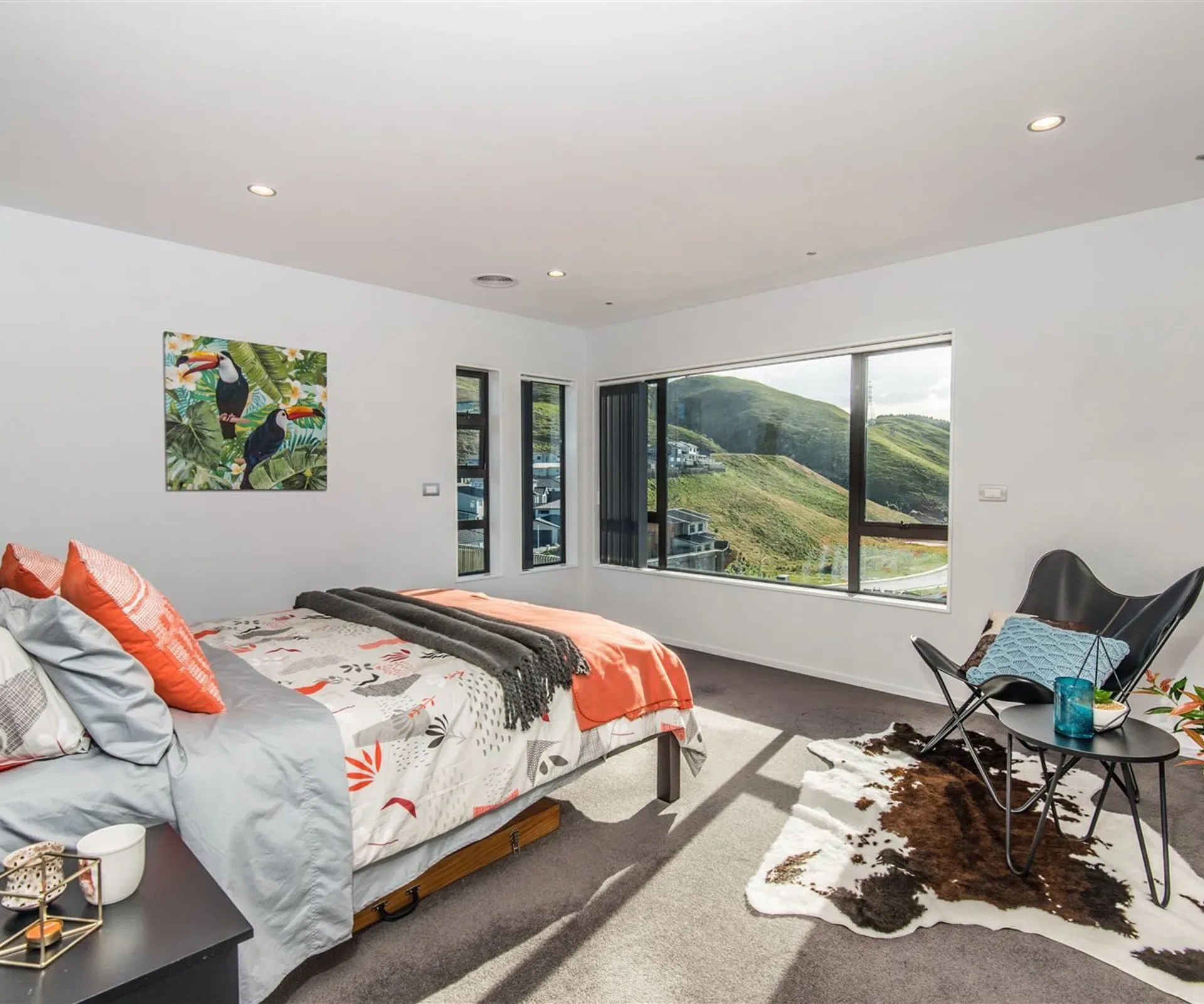 The main bedroom — the bed under a corner run of windows onto the hills, a cowhide rug and a black sling chair beside it, in hard morning light.