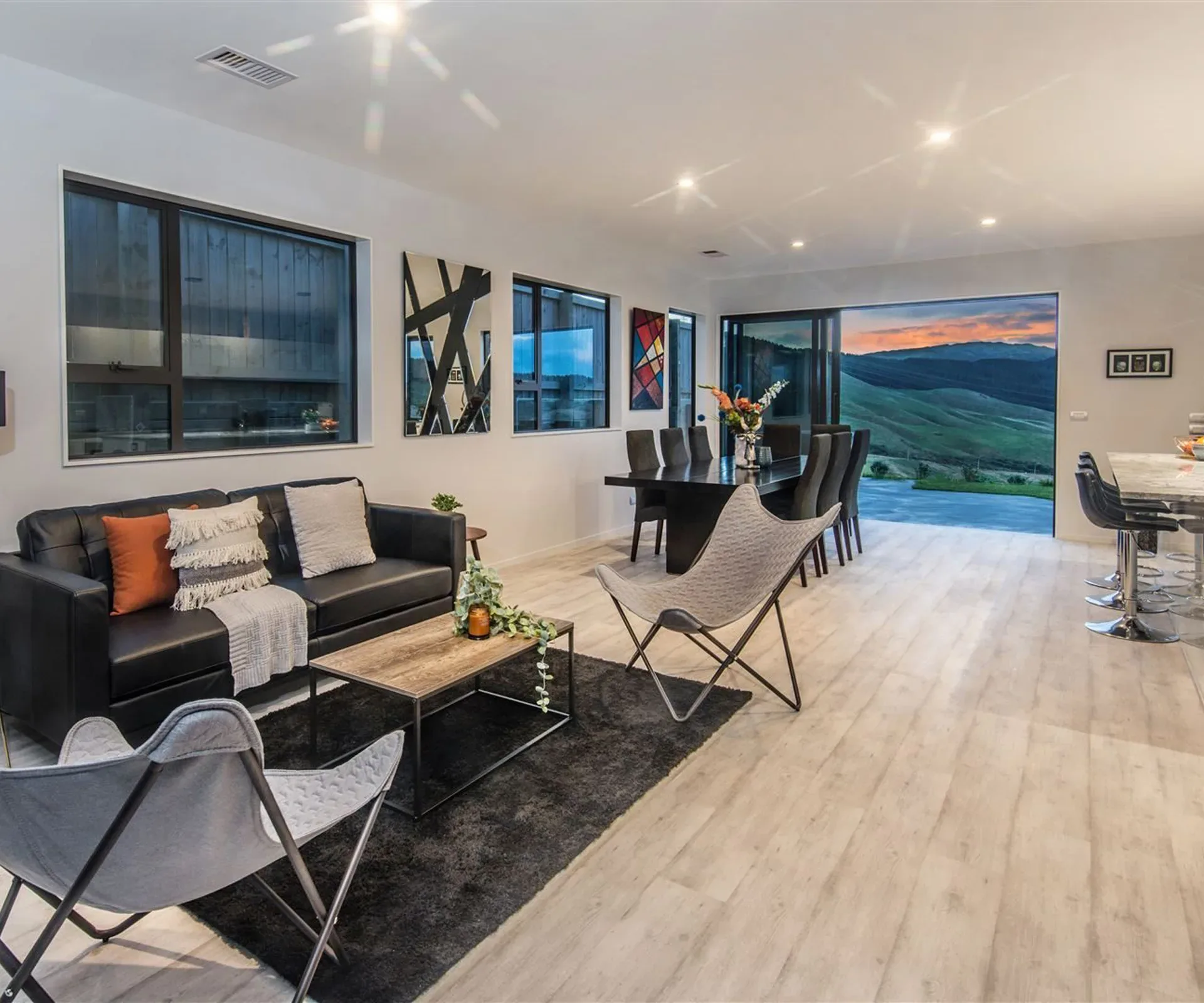The open-plan living area — sofa and sling chairs on a dark rug in the foreground, the dining table and kitchen bar beyond, and the valley framed in the full-height slider at the end of the room.