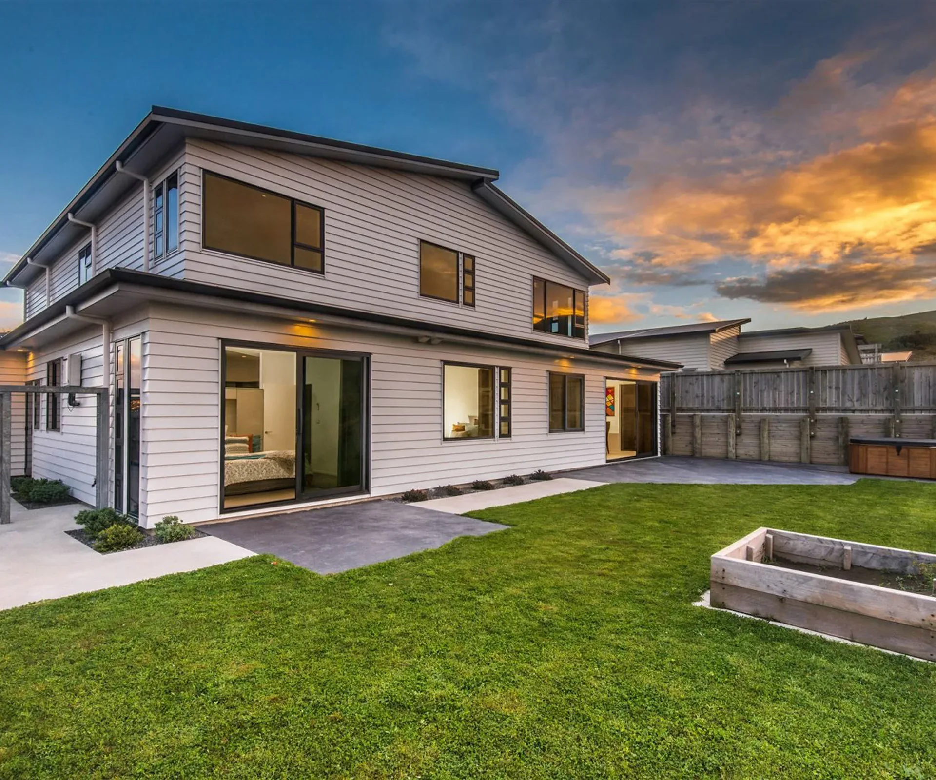 The rear of the house from the lawn at dusk — the sliders and upstairs windows lit from inside, a spa set into the deck against the timber fence, and a raised timber garden bed on the grass.
