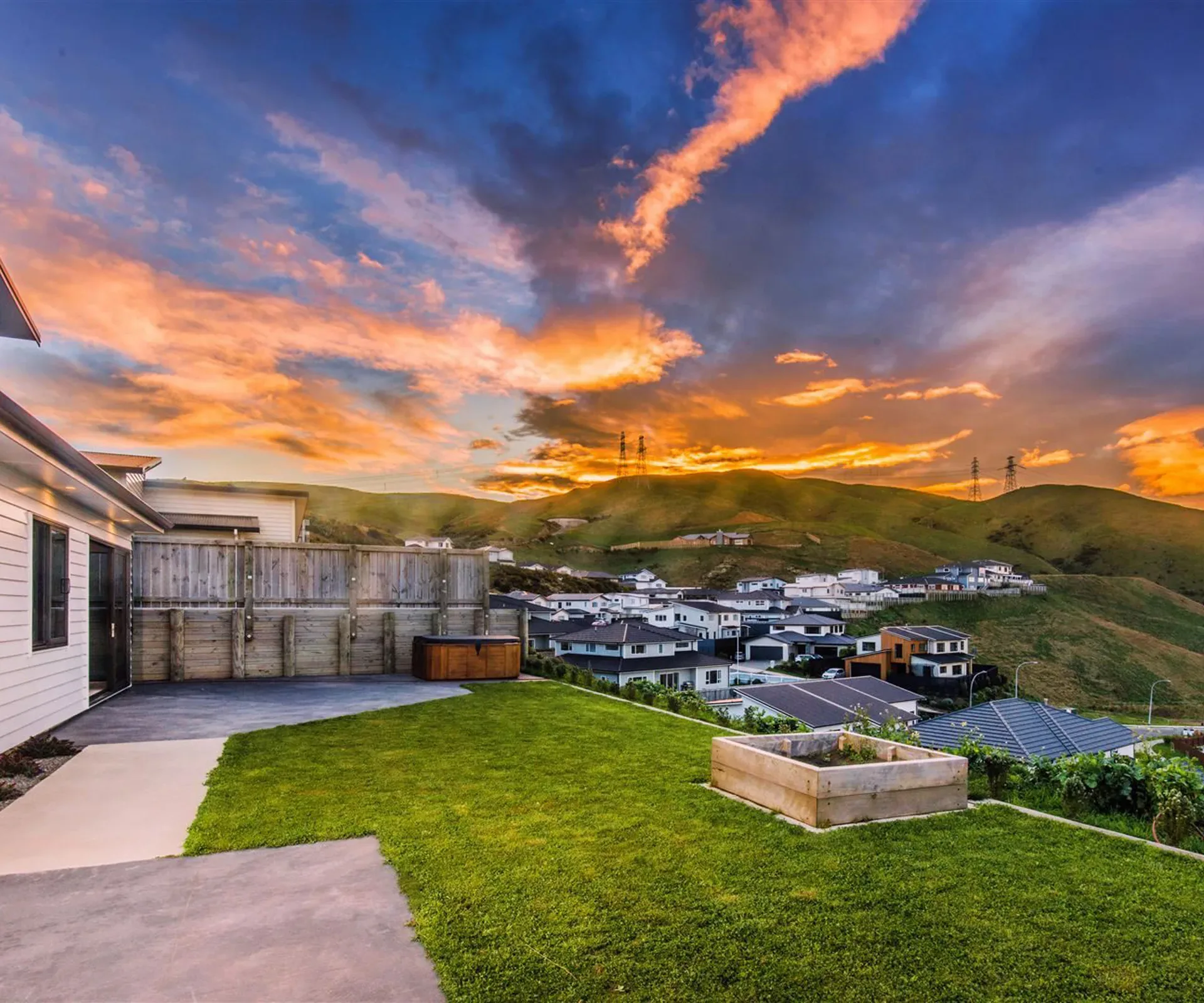The rear yard at sunset — the timber retaining wall and boundary fence along one side, the spa on the paving, a raised timber garden bed on the lawn, and the suburb and green hills falling away below.