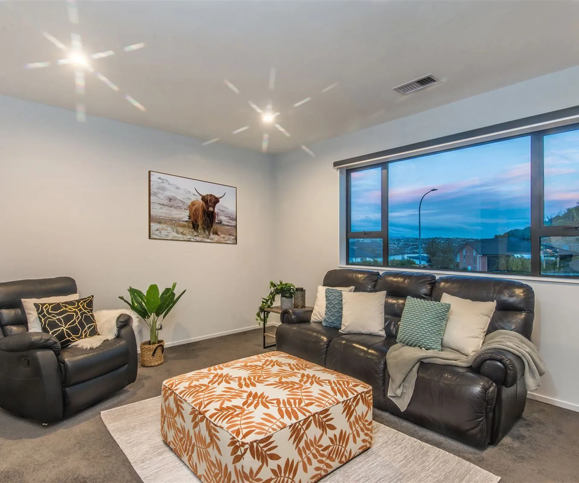 The second living room — a black leather recliner suite around a patterned ottoman, and a long window running the wall behind them onto the town and hills at dusk.