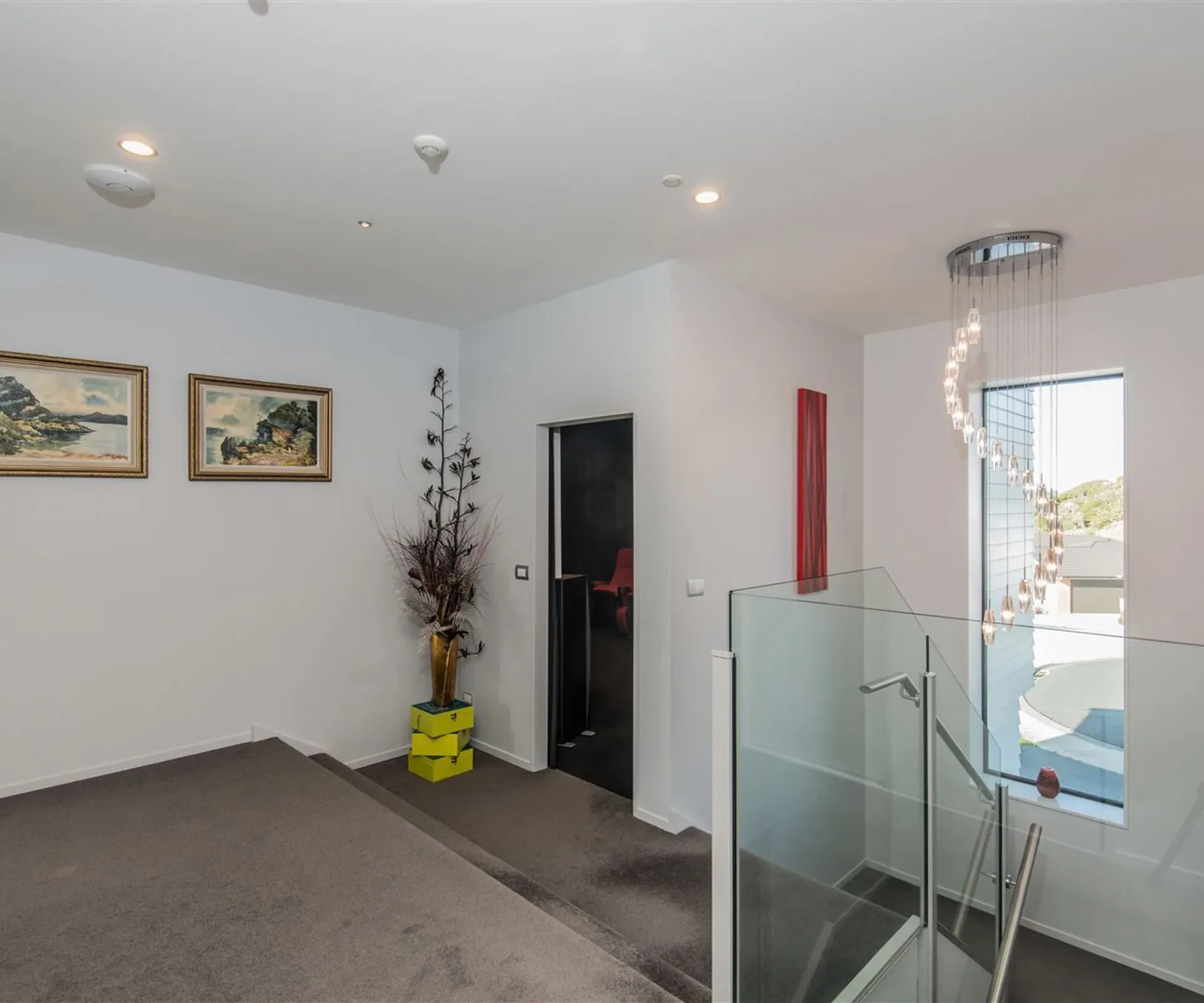 The upstairs landing — a glass balustrade at the head of the stair, a cluster of pendants hung down the stairwell window, and an open doorway into the dark cinema with one of its red chairs showing.