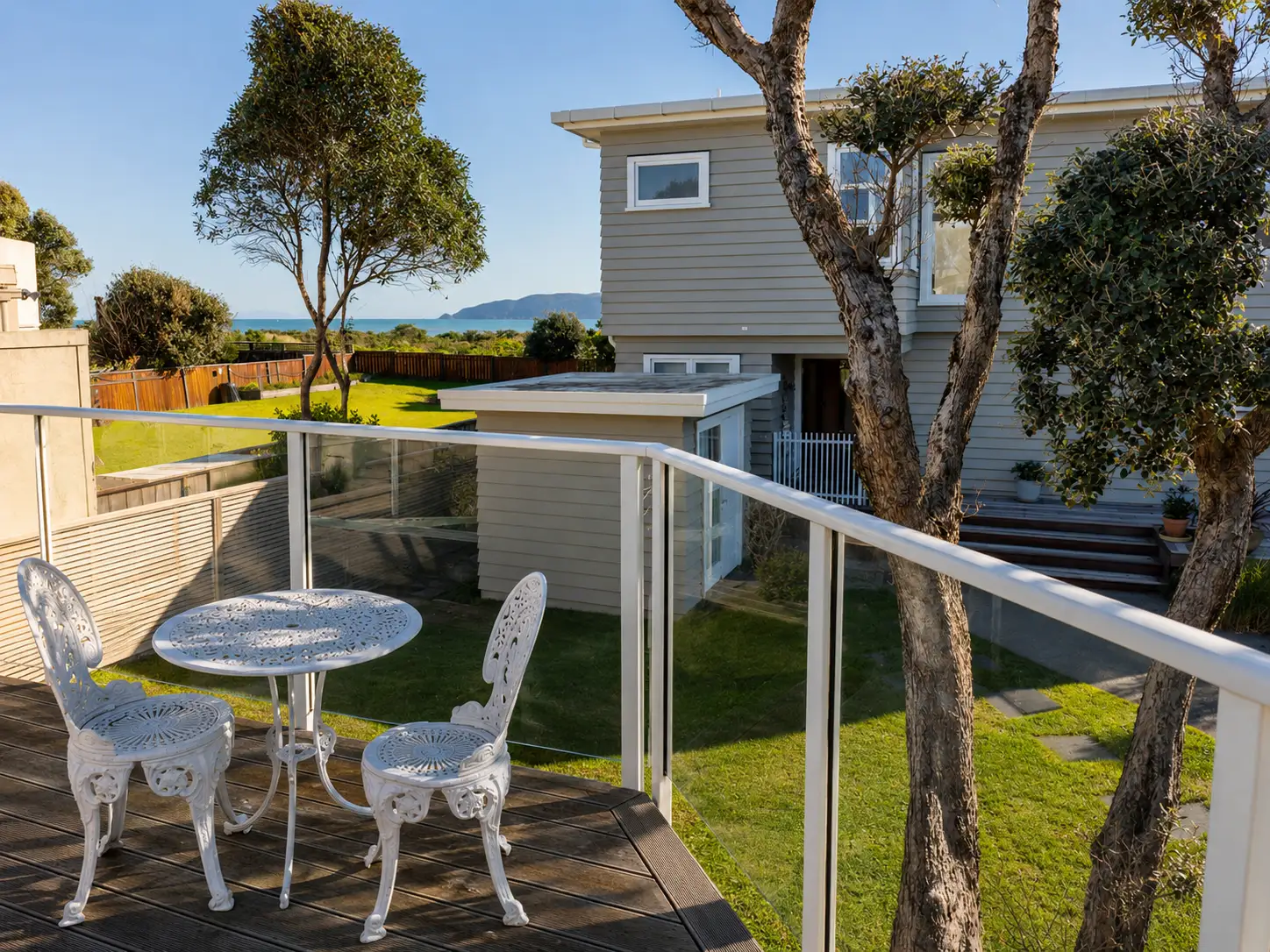 Manly Street — the kwila deck off the guest quarters, glass balustrade, table and chairs, with the lawn below and the sea beyond