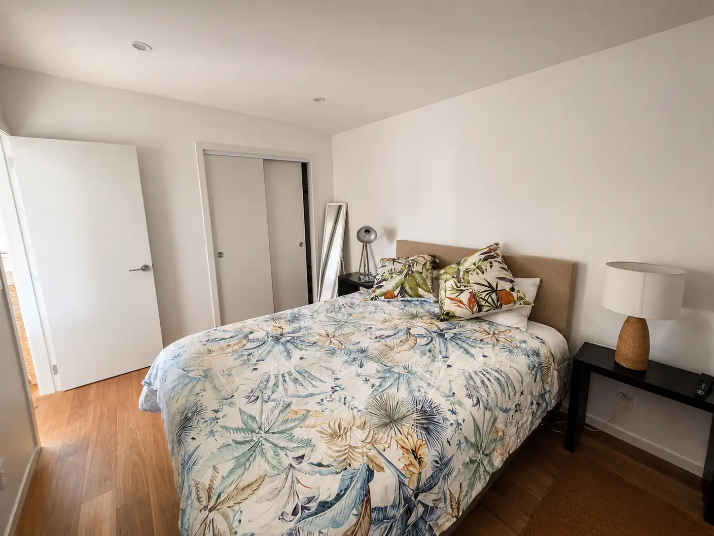 The bedroom from the doorway — the bed under a leaf-print cover, the wardrobe doors beside it and a mirror leaning by the bedhead.