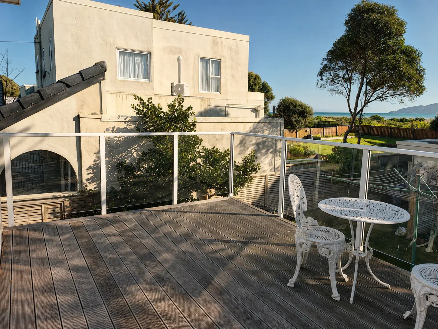 The deck looking out past the glass balustrade — the neighbouring house on one side, planting on the other, and the sea along the horizon.