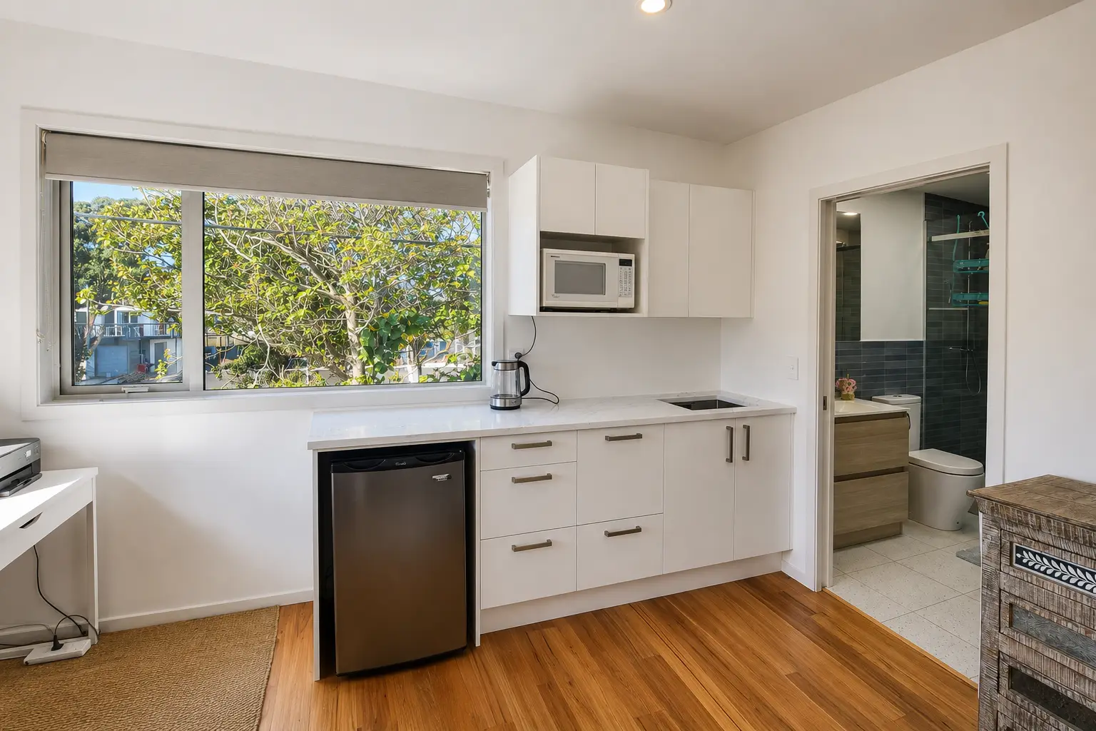 The kitchenette — white cabinetry along one wall with a microwave over the bench, an undercounter fridge, and the bathroom door open beyond it.