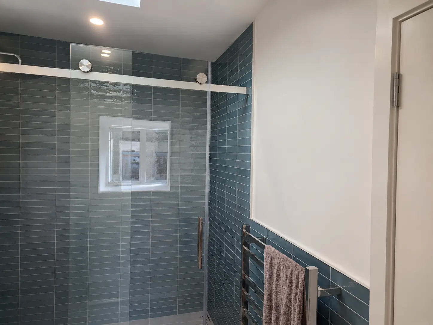 The shower behind its sliding glass — dark blue-green tile to the sill line, a small window in the return, and a heated towel rail on the wall alongside.