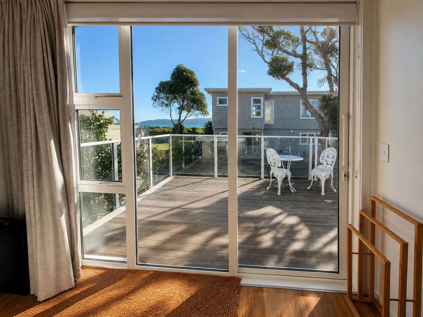 The slider onto the deck seen from inside, curtains drawn back, the table and chairs and the trees beyond the glass.