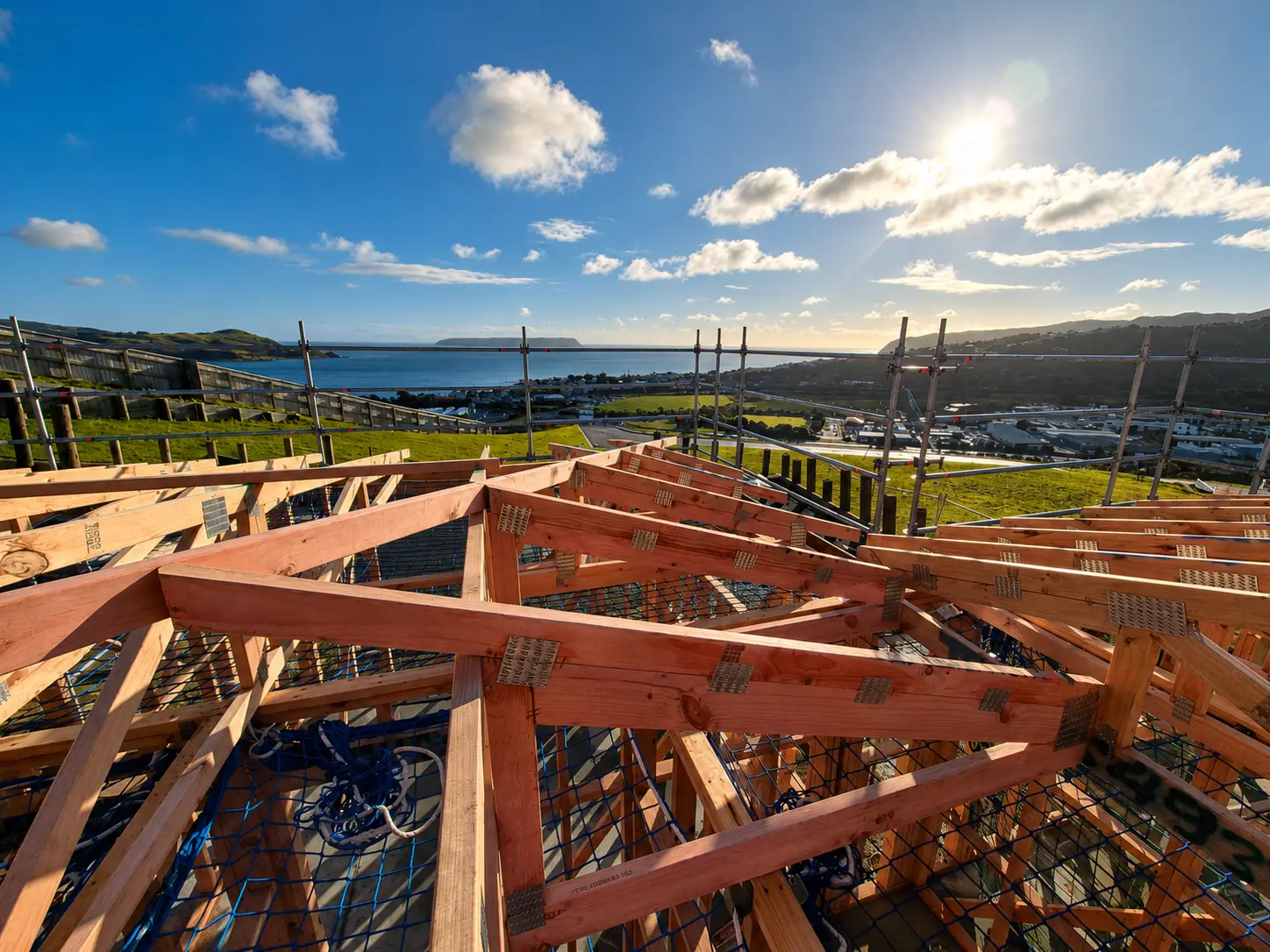 Along the ridge with the roof structure set out and braced, nail plates catching the sun, and the paddocks, road and water dropping away below the scaffold.