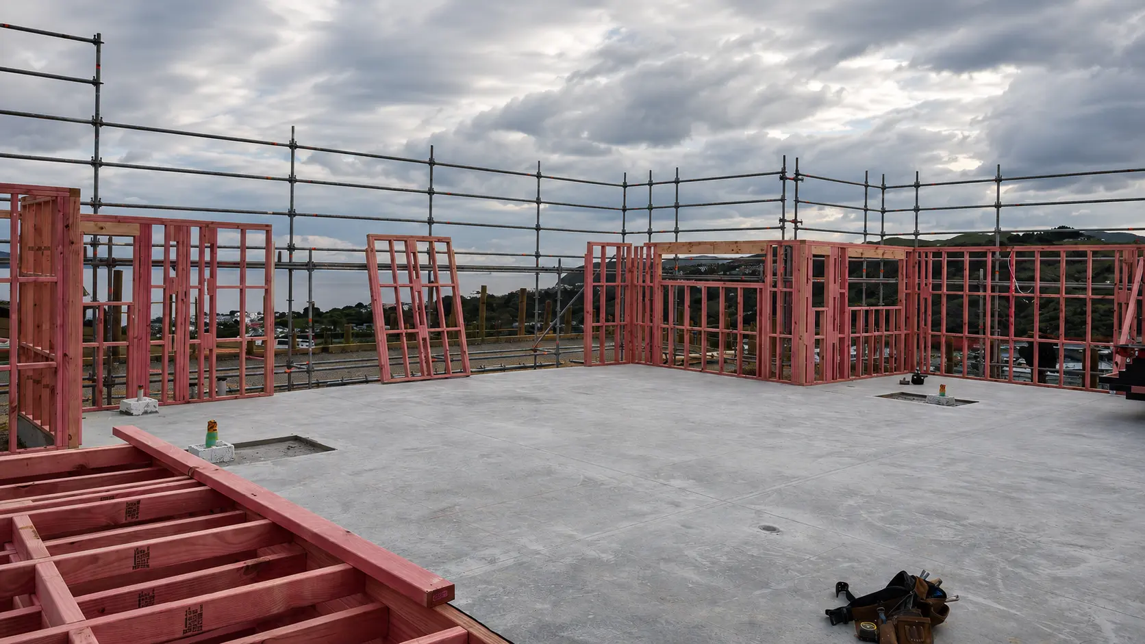 The first wall frames standing on the slab under an overcast sky — one bay braced off the floor, another laid flat against it, and the edge-protection scaffold ringing the platform.
