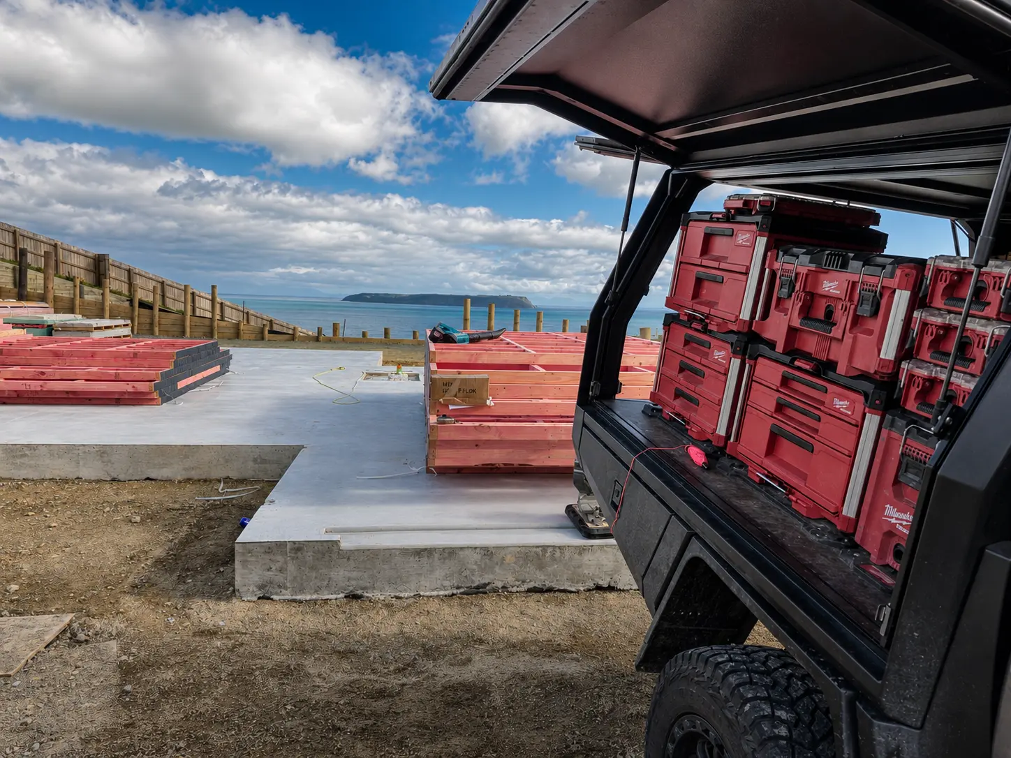 The slab from the driveway with the work ute backed up to it, canopy open on a bank of tool cases, and the stacked frames and pole retaining wall running away toward the water.