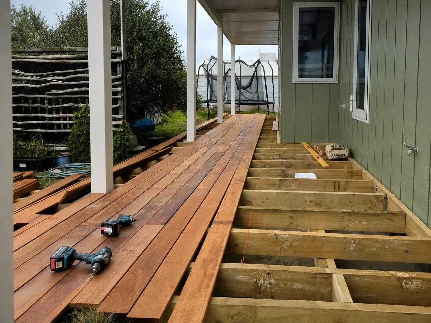 The boards going down — laid kwila on the left running to a string line, the open joists still showing on the right, and two cordless drills resting on the finished half.