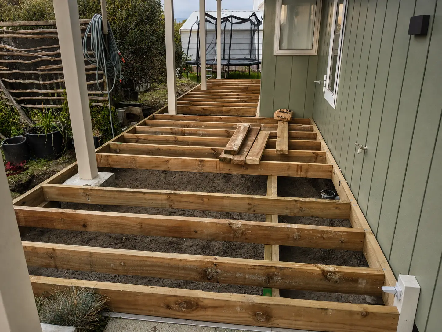 The substructure complete — treated bearers and joists framed out over the ground between the house wall and the boundary, boxed around the veranda posts and running away to the step at the far end.