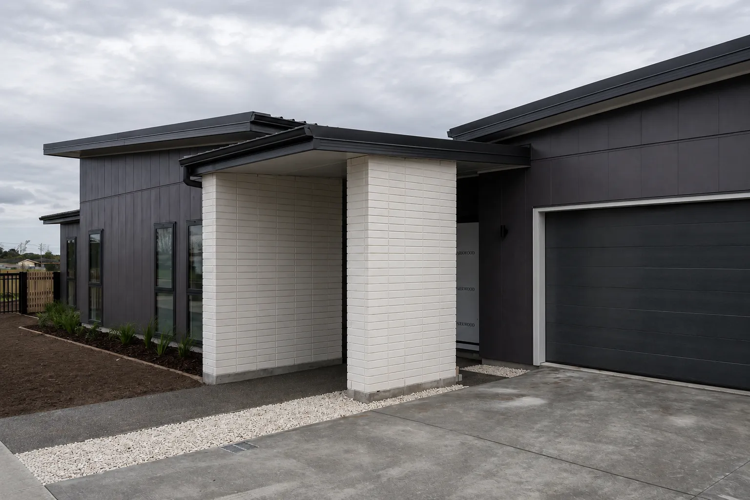 The entry portico close up — the pale brick piers carrying the roof over the front door, the dark boards and window joinery running away to the left, and a white shingle strip along the base of the wall.