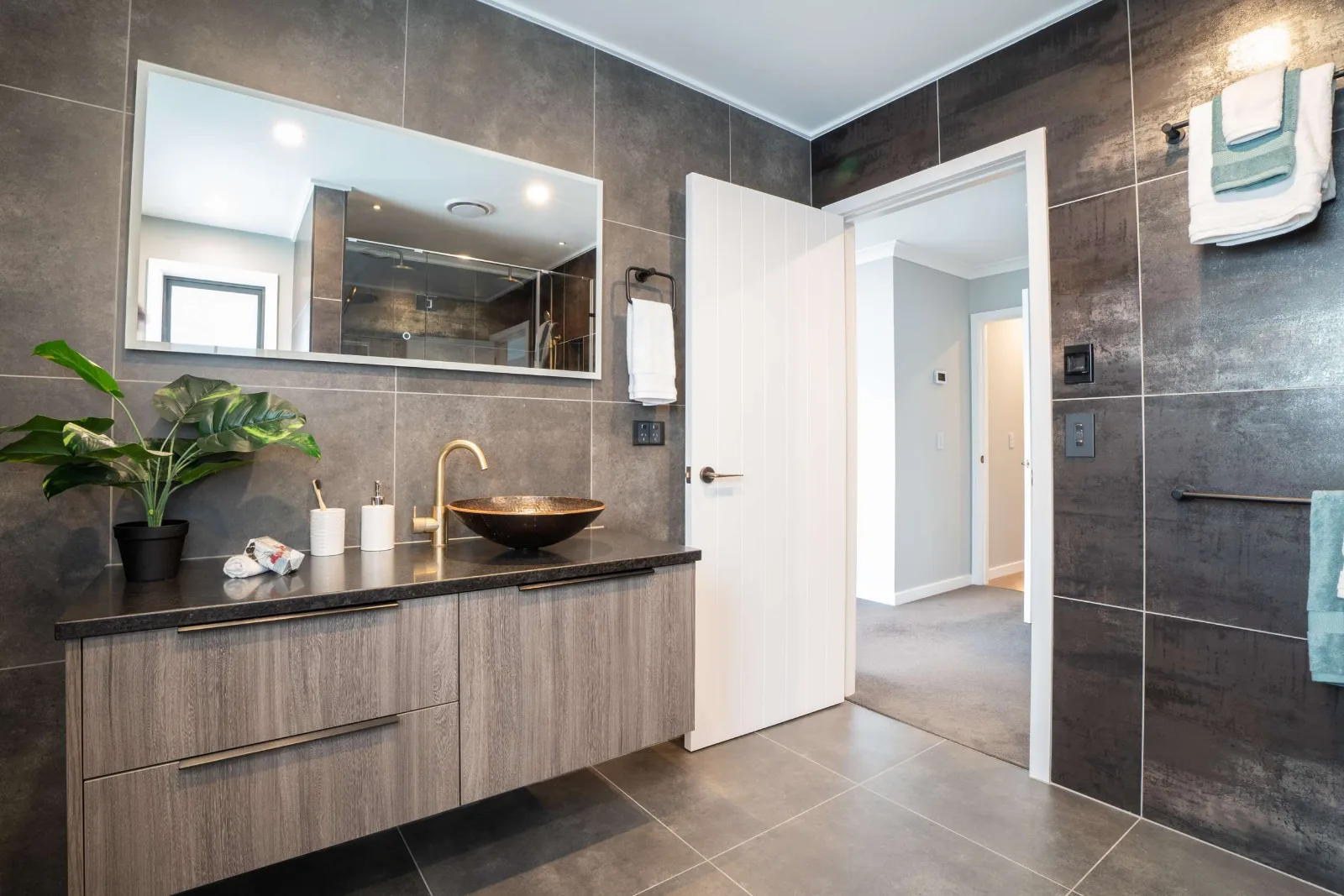 A bathroom with a hammered vessel basin on a dark stone top, brass tap and a long mirror above it, and the door standing open to the carpeted hall.