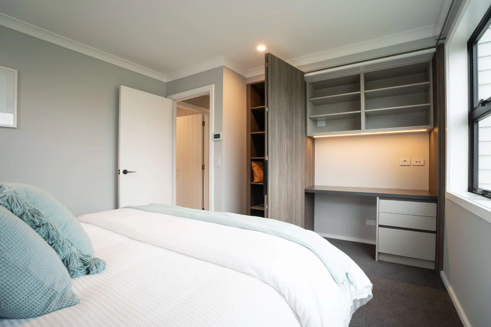 A bedroom with a built-in desk and shelving alongside the wardrobe, the shelf lit from beneath, and the door standing open to the hall.