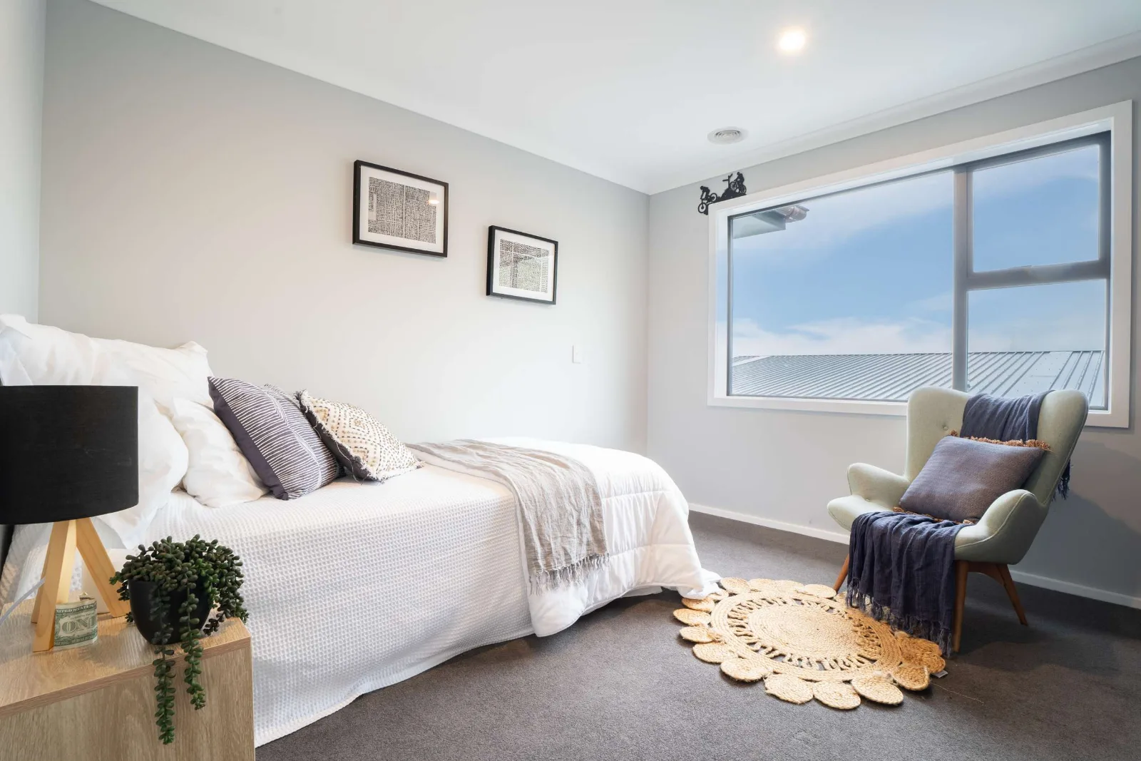 A bedroom with two framed prints over the bed, a window onto the neighbouring roofline and sky, and a pale armchair on a round jute rug.