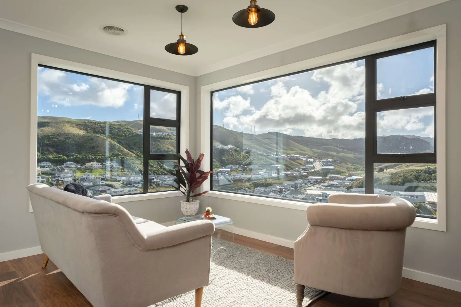 The corner sitting room — two walls of glass meeting at the corner over the valley, the houses below and the hills beyond, with brass pendants hung in front of them.