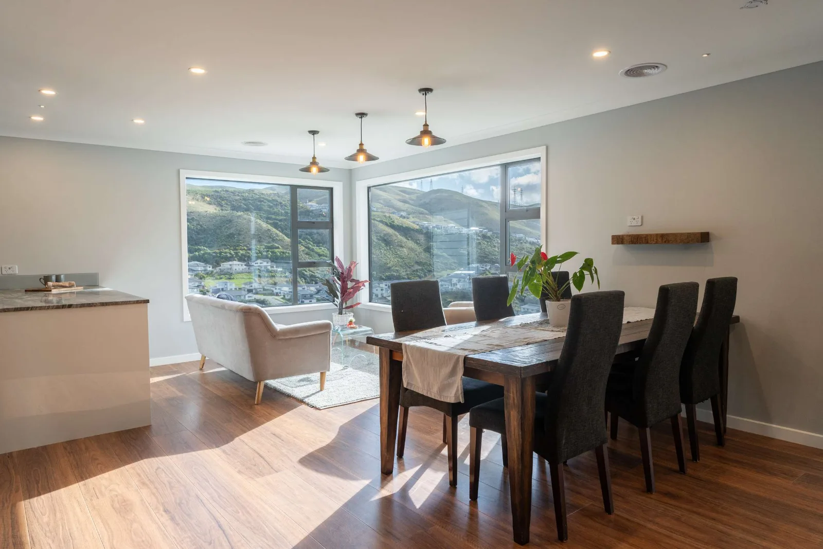 The dining end — a long table under three brass pendants, the kitchen bench running away to the left and the valley filling the windows behind.