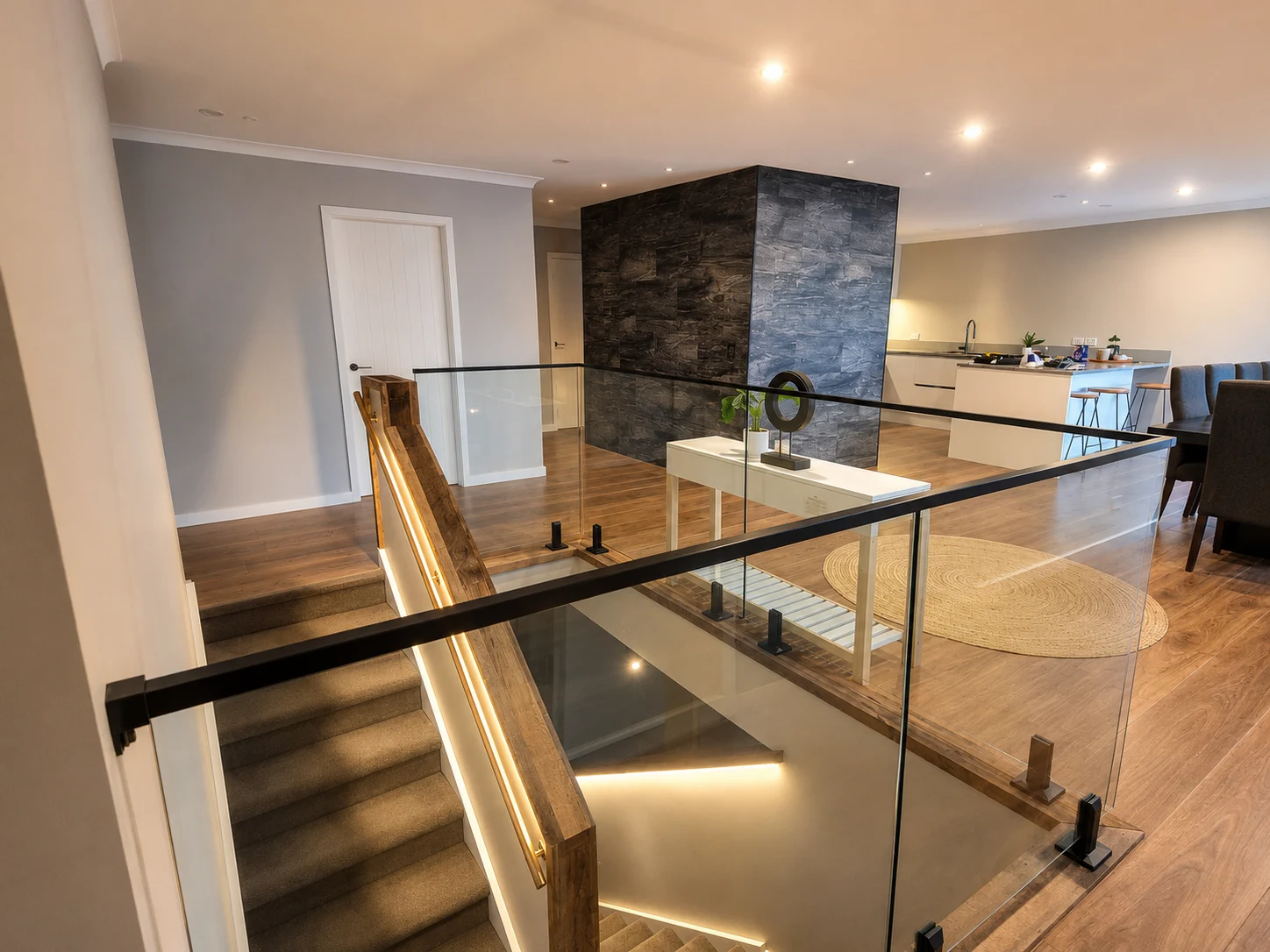 The feature stair seen from the upper landing — timber handrail with the LED run set into it, glass balustrade around the void, and the dark stone-clad column, kitchen and dining beyond.