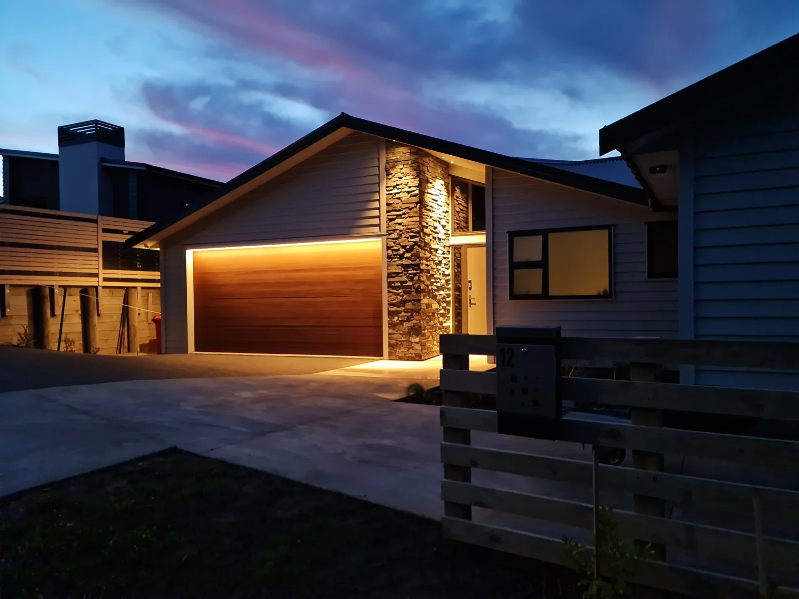 The garage and entry at dusk — the timber door lit along its head from a concealed strip, the schist column uplit the full height of the gable beside the entry door, and the numbered letterbox standing at the slatted fence.
