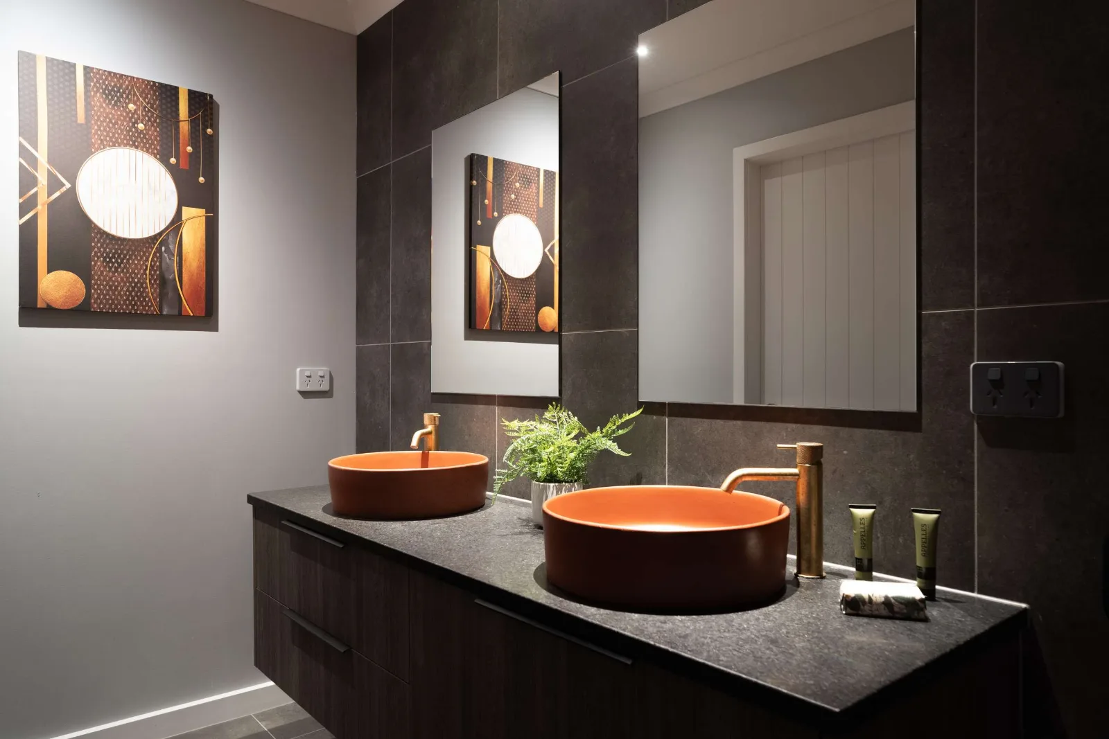 Two hammered copper basins on a dark stone top — brass tapware, dark tile to the ceiling, a backlit mirror and a copper-toned canvas on the end wall.