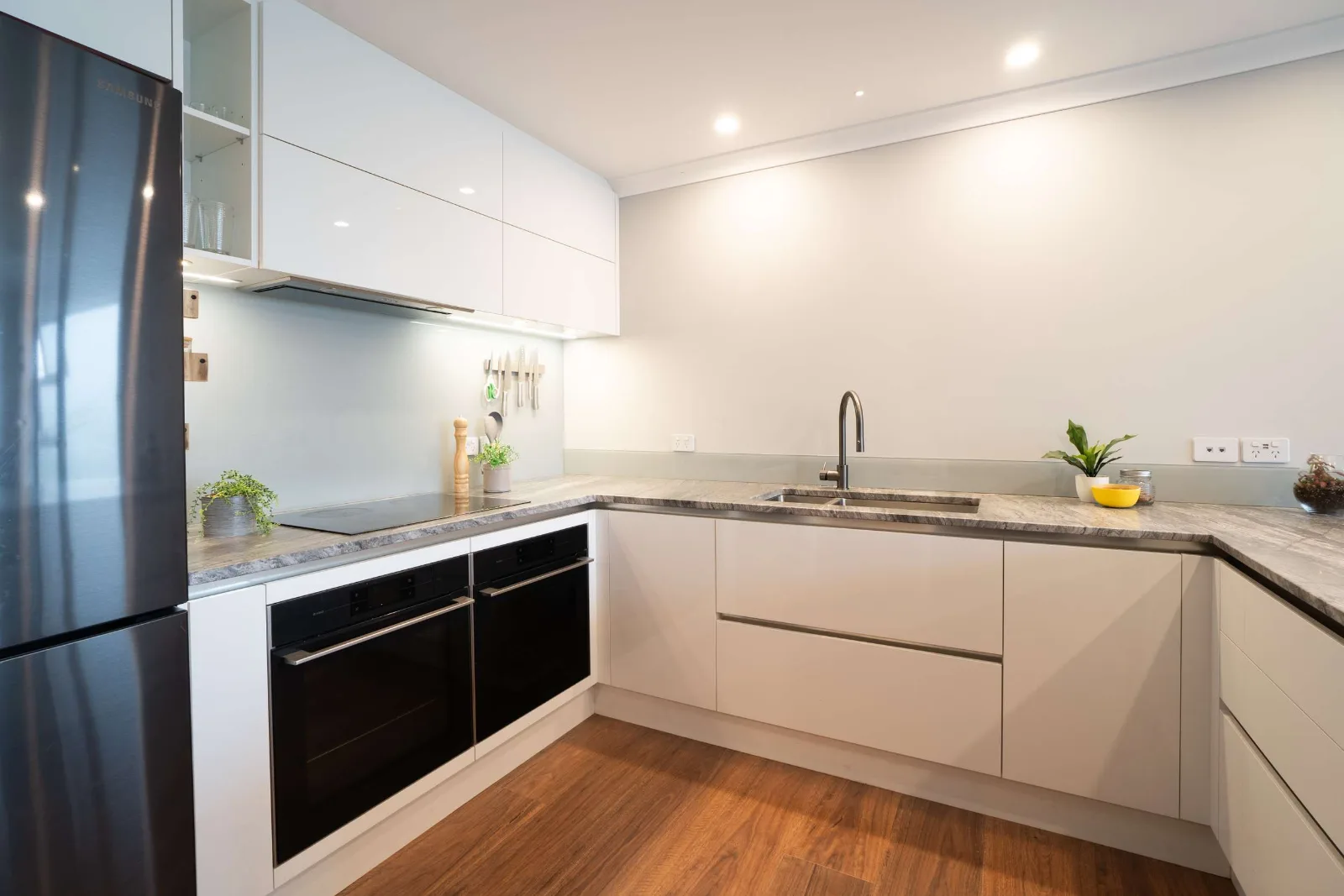 The same kitchen along its length — the induction hob and ovens on the left, the sink set into the run of bench, and the timber floor turning the corner.