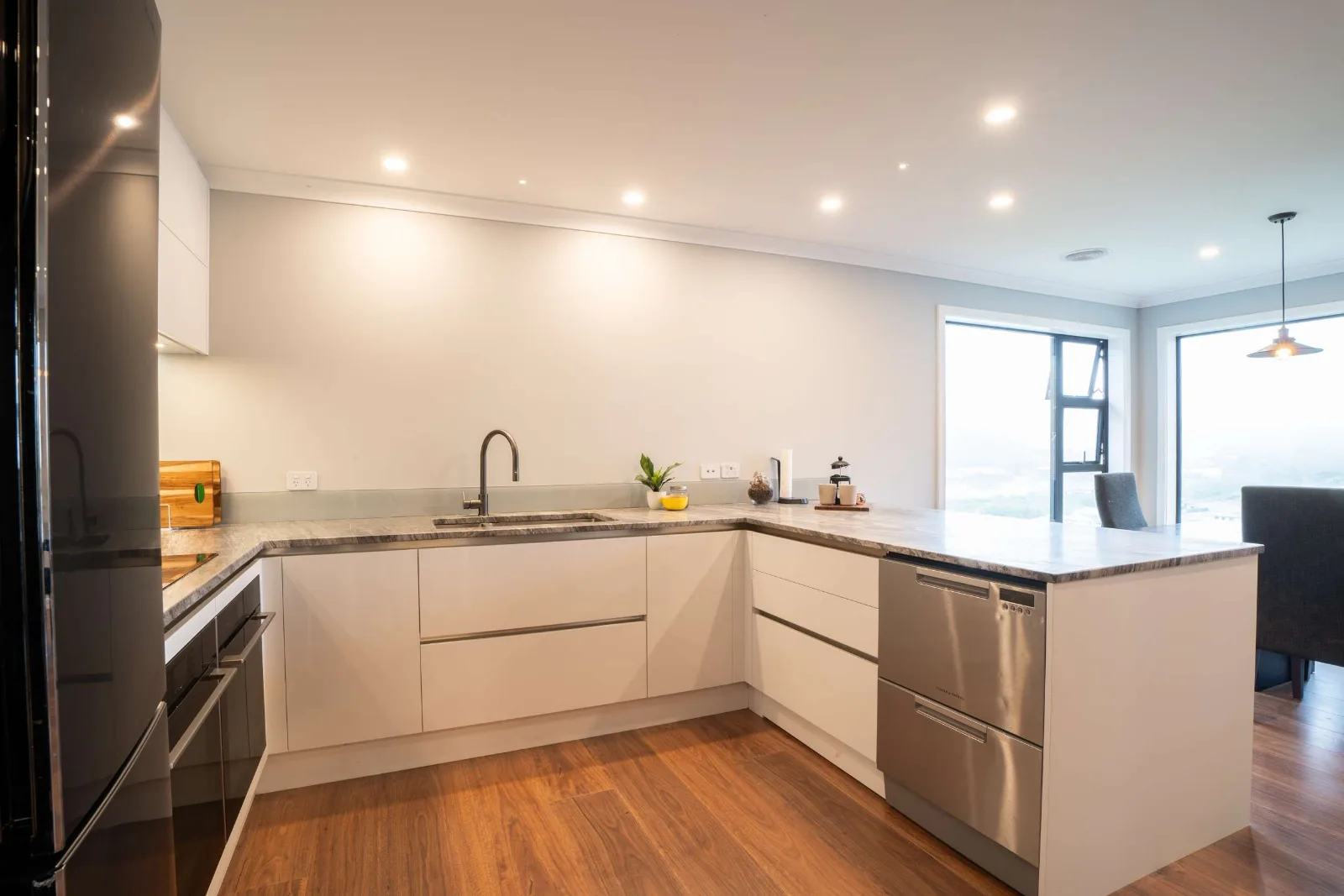 The kitchen from the dining end — the full run of white cabinetry, the dish drawers set into the peninsula, and the windows onto the valley beyond it.