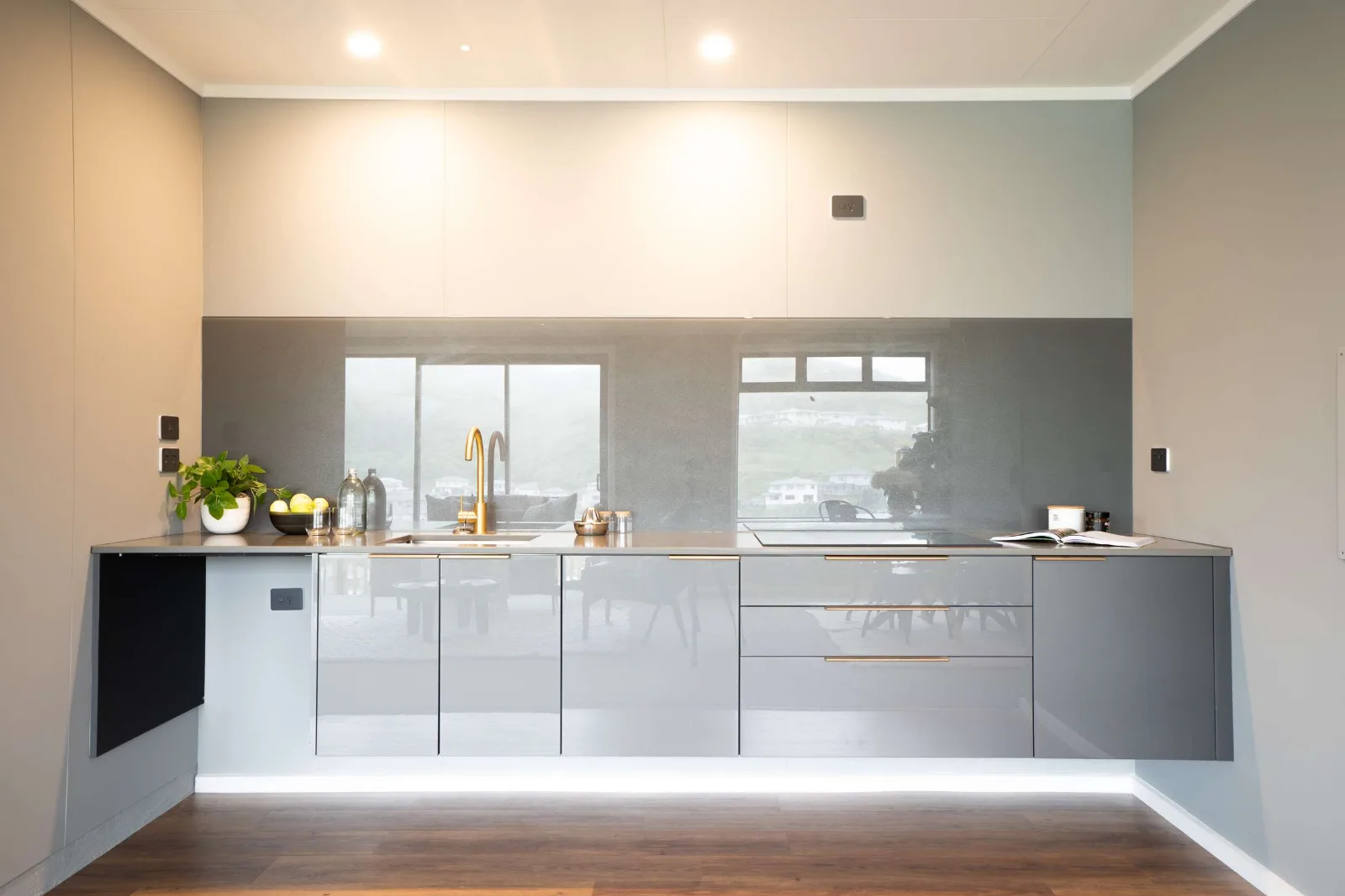 A kitchen run in grey gloss with brass handles and a brass tap, a mirrored splashback carrying the windows back into the room, and the cabinetry lifted clear of the floor on a lit shadow line.