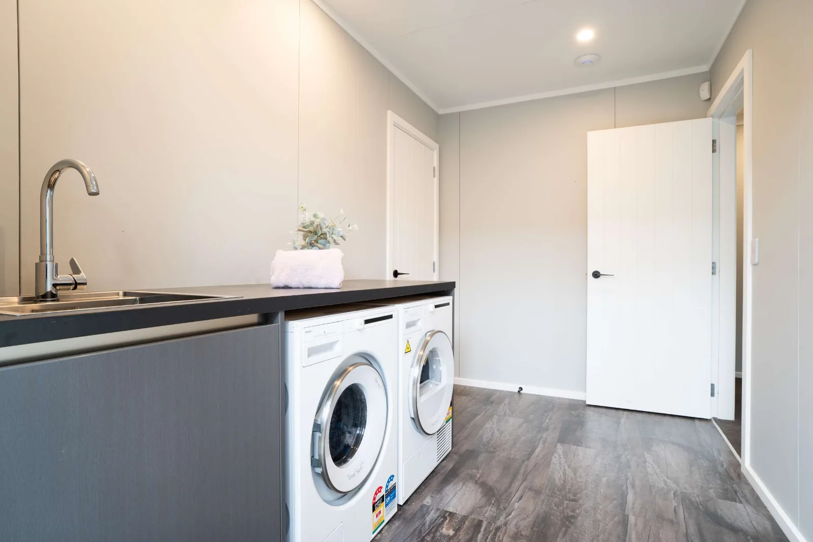 The laundry — a stainless tub set into a dark benchtop with the washer and dryer under it, and two doors off the far end.
