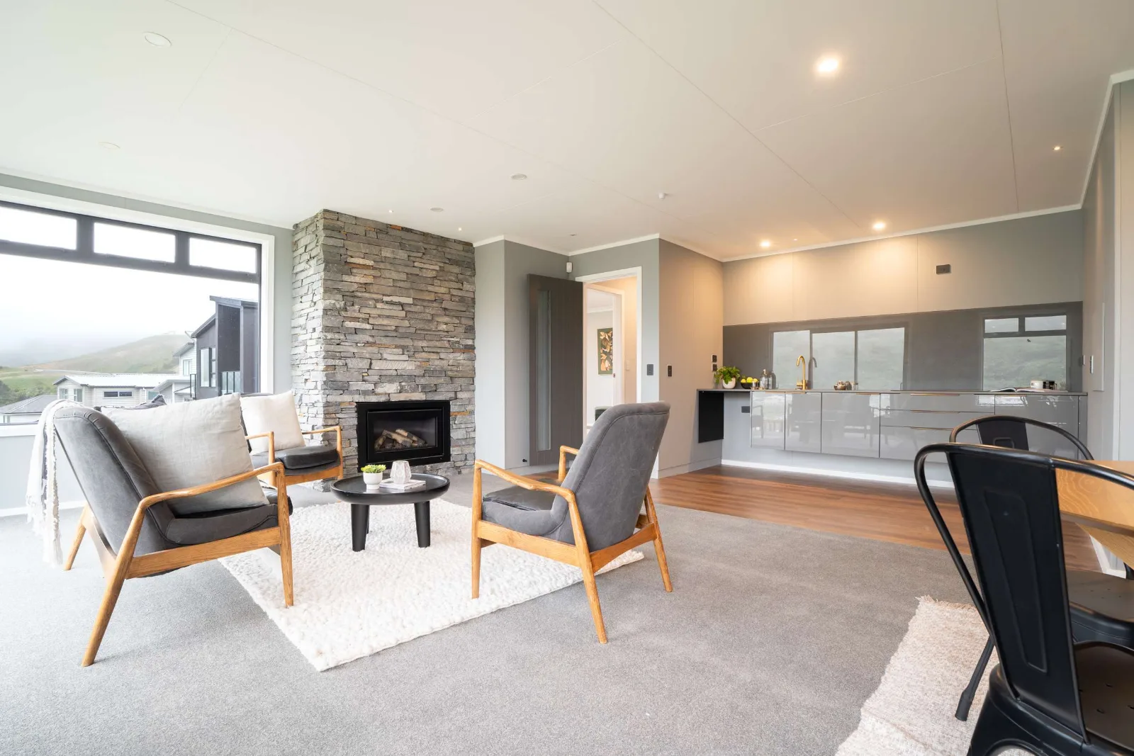 The living area — the schist chimney breast with the gas fire alight, two timber-framed armchairs on a wool rug, and the kitchen and dining beyond it under the same ceiling.