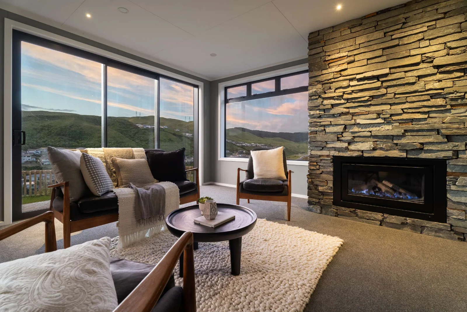 The living area with the fire alight — the schist chimney breast running the full height of the wall, chairs and a low round table on a wool rug, and the hills and sunset filling the corner glazing.