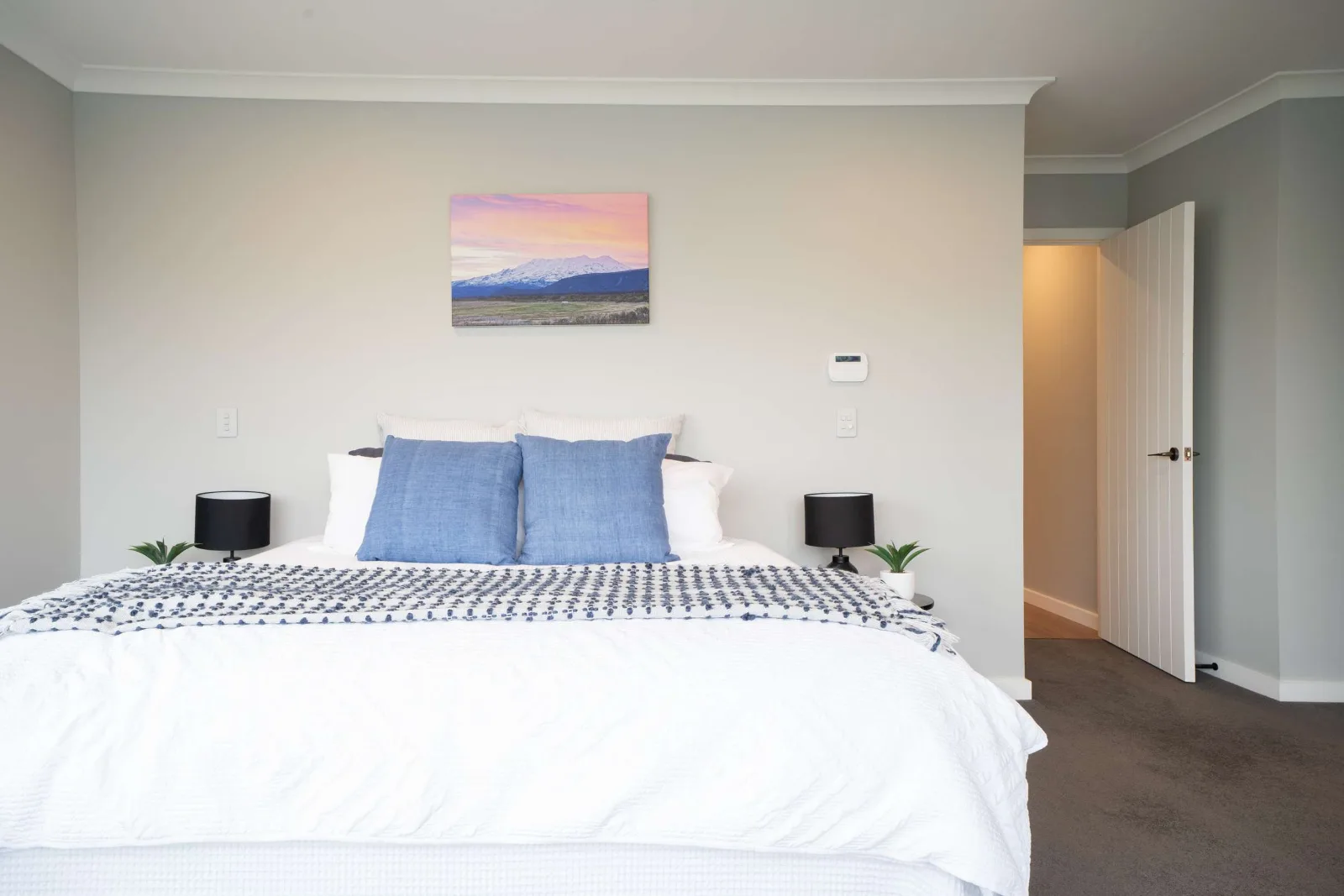 The main bedroom from the foot of the bed — a mountain canvas centred over the bedhead, a lamp to each side, and the door open to the hall.