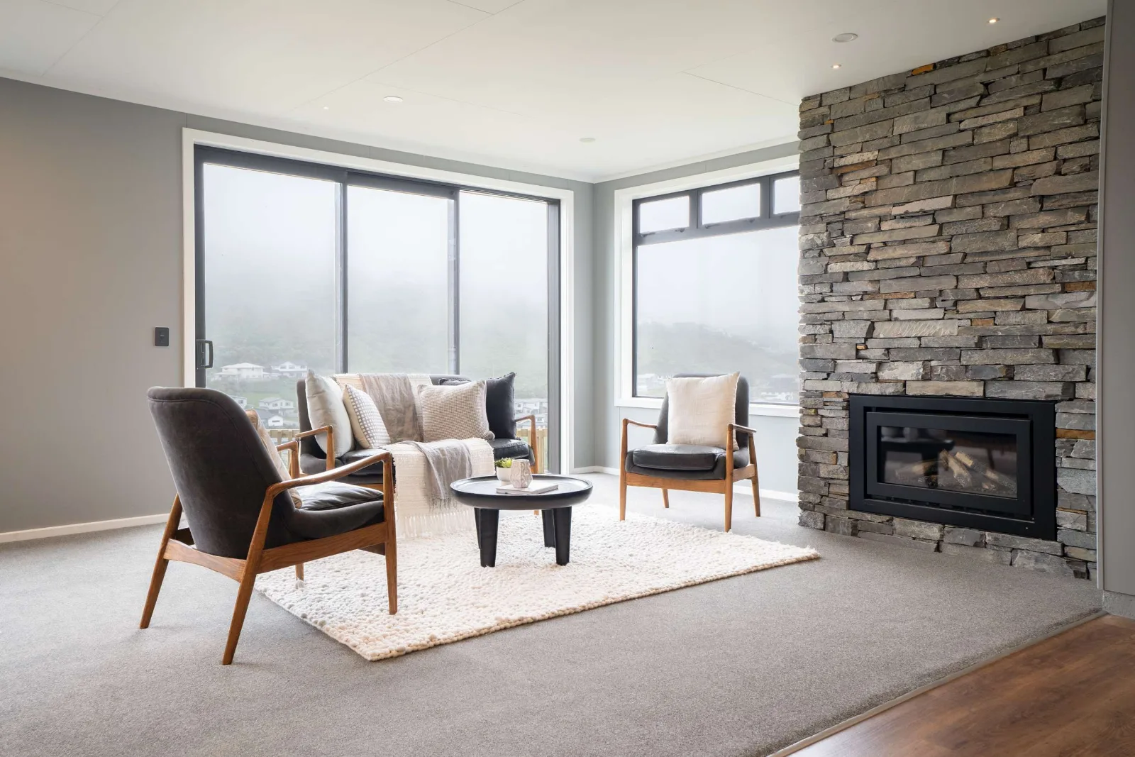 The same room on a still, foggy morning — the corner glazing white with cloud, the schist chimney breast beside it, and the timber floor of the dining end at the right.
