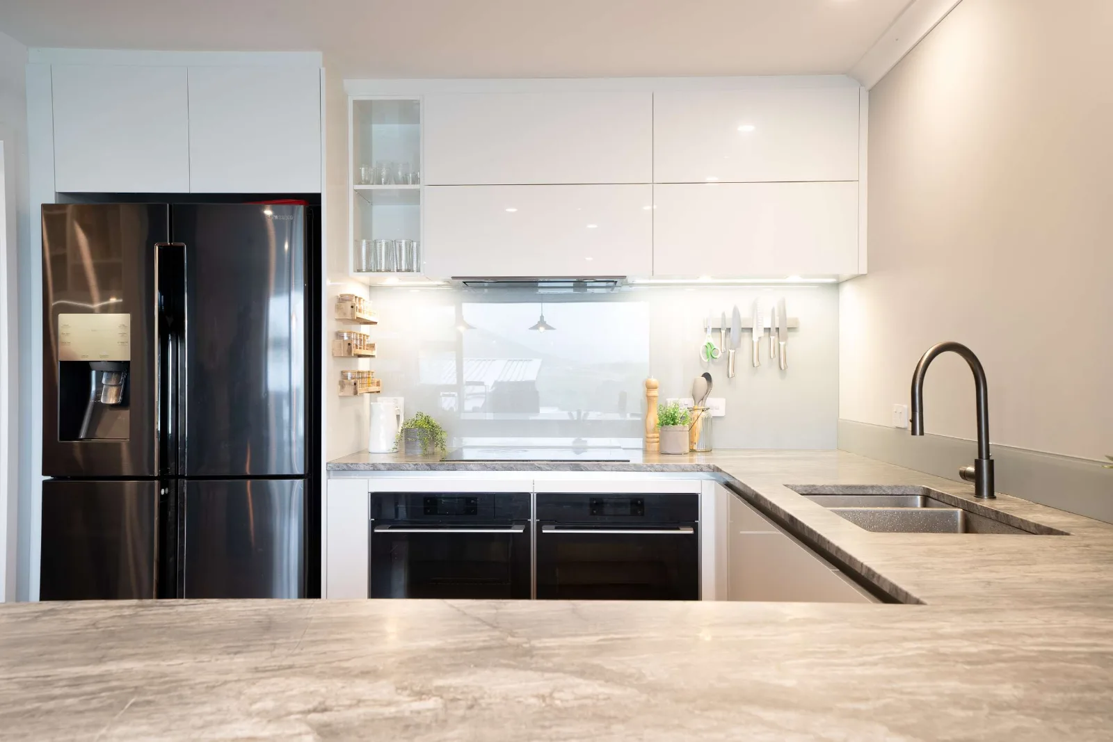 The second kitchen — white gloss cabinetry with a glass splashback and a window through it, the wall ovens under the bench, and the stone-topped peninsula in the foreground.