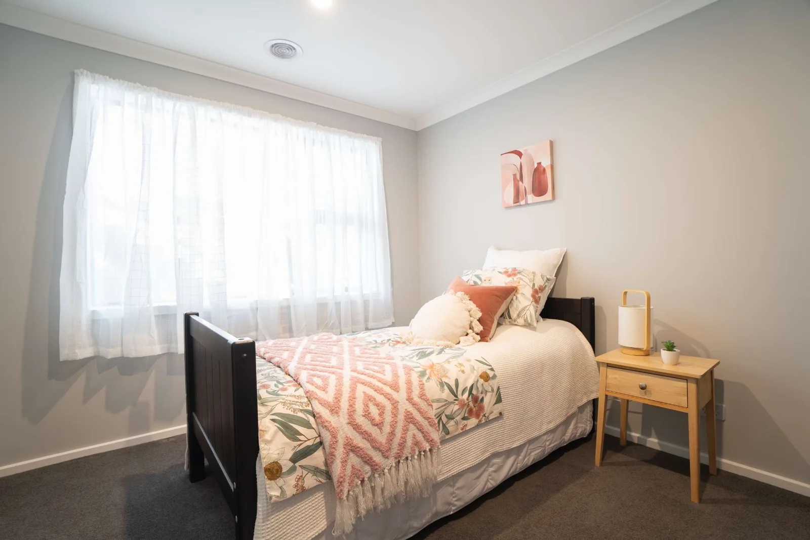 A single bedroom — the bed against the wall under a framed print, a sheer-curtained window beside it and a timber side table.