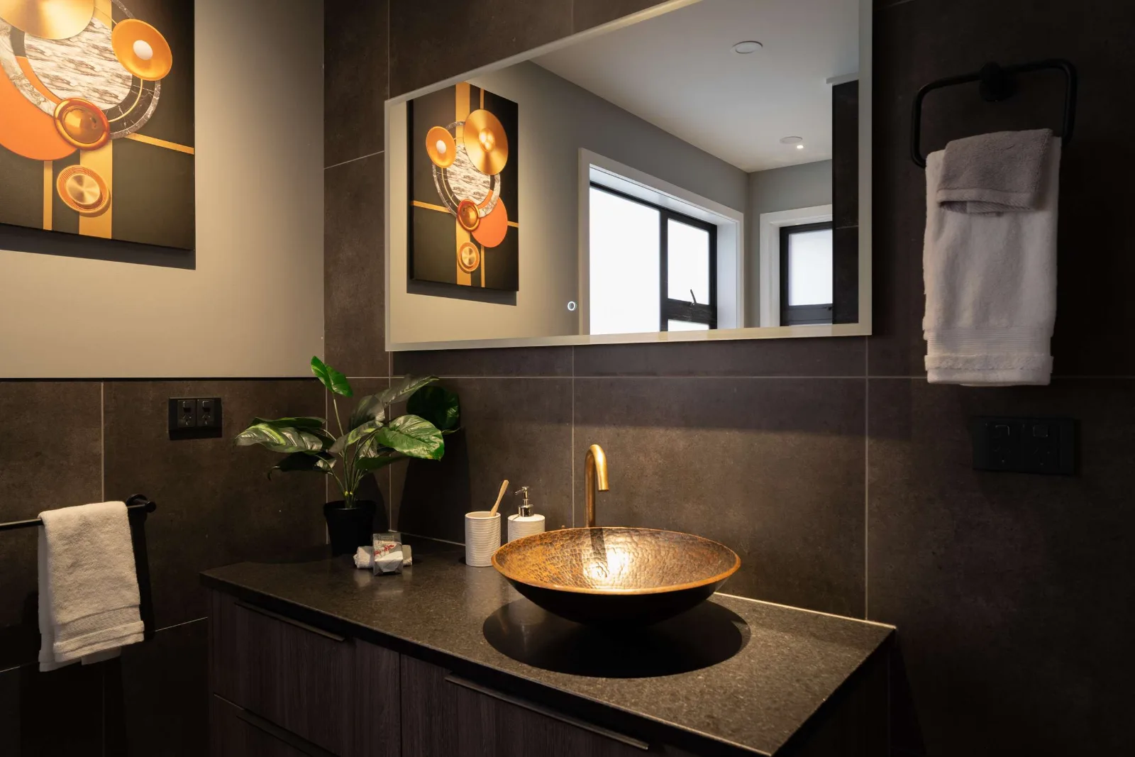A single hammered copper basin on a dark stone top, brass tap and backlit mirror, with the gold-and-orange canvas from the wall repeated in the glass.
