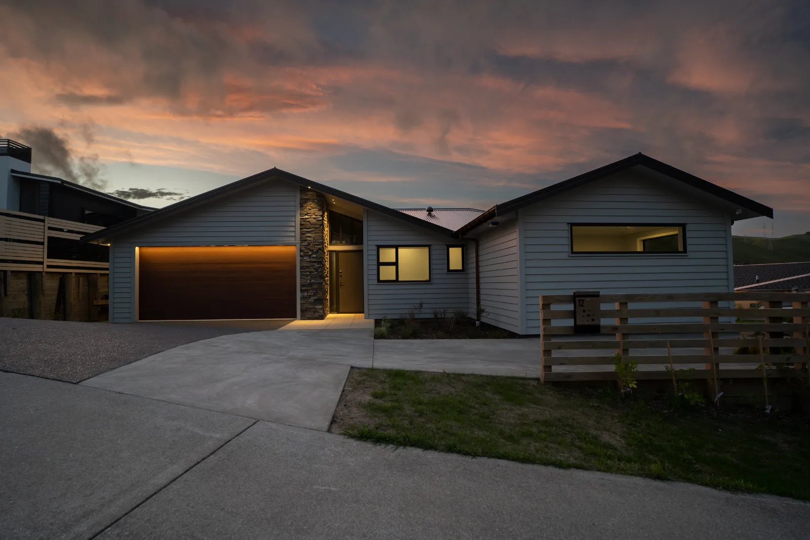 The street front under a pink sunset sky — both gables, the timber garage door lit from within its opening, the schist column standing the full height of the entry, and the numbered letterbox on the slatted fence.